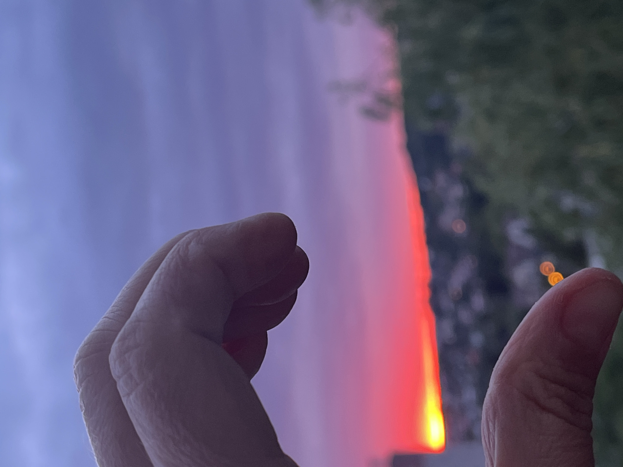 An over-the-shoulder, close-up shot captures a hand against a blurred twilight sky. The hand is positioned in the upper left of the frame, with fingers gently curved. In the background, a vibrant sunset paints the horizon with streaks of red and yellow, transitioning to shades of purple and blue in the sky. Below the sunset, a blurred cityscape with scattered lights is visible, nestled on a gentle incline. The scene conveys a serene, atmospheric moment as day transitions into night.