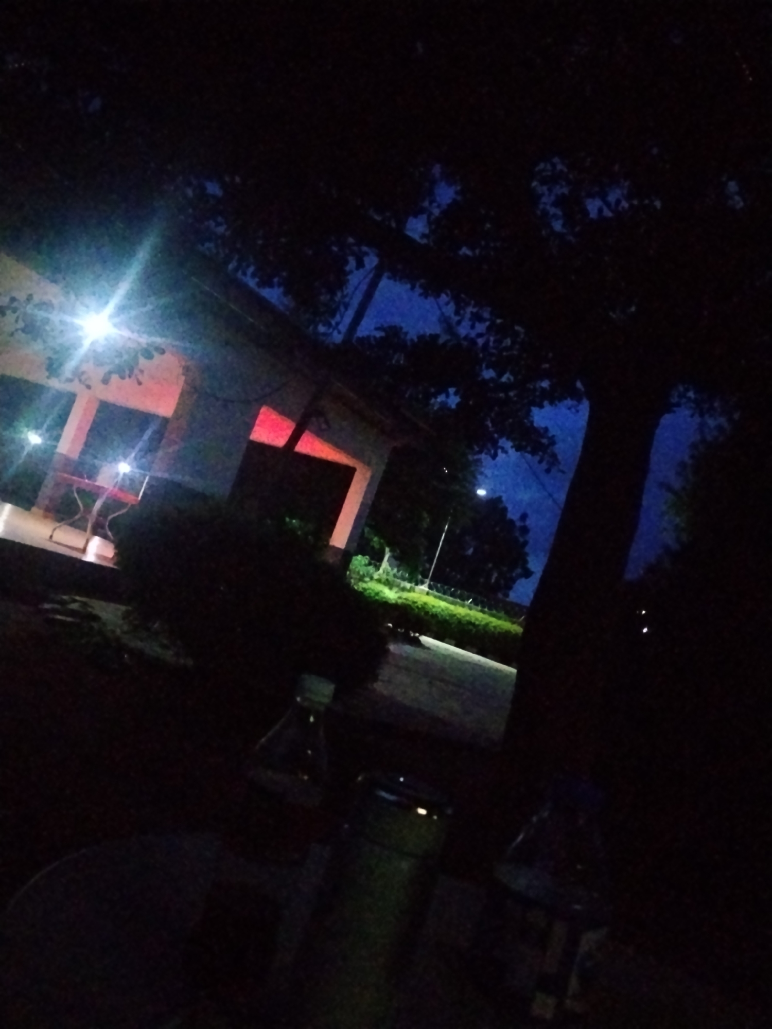 The image depicts a scene at dusk or night in Mangu Town, Nigeria. The foreground is dark and slightly out of focus, with a partially visible plastic bottle and a cylindrical object that could be a thermos or a bottle. In the mid-ground, a building or structure with white walls and a covered porch is illuminated by lights. Red light is visible inside one of the rooms of the structure. There is a red table and chairs on the porch. A tree with dark foliage dominates the right side of the image, silhouetted against a dark blue sky. Further in the background, a street light and a hedge are visible, along with more trees and the dark silhouette of what could be a vehicle. The overall atmosphere is quiet and subdued, suggesting a relaxed evening setting. The time of day is clearly night or late evening due to the darkness and artificial lighting. There are no visible text elements in the image.