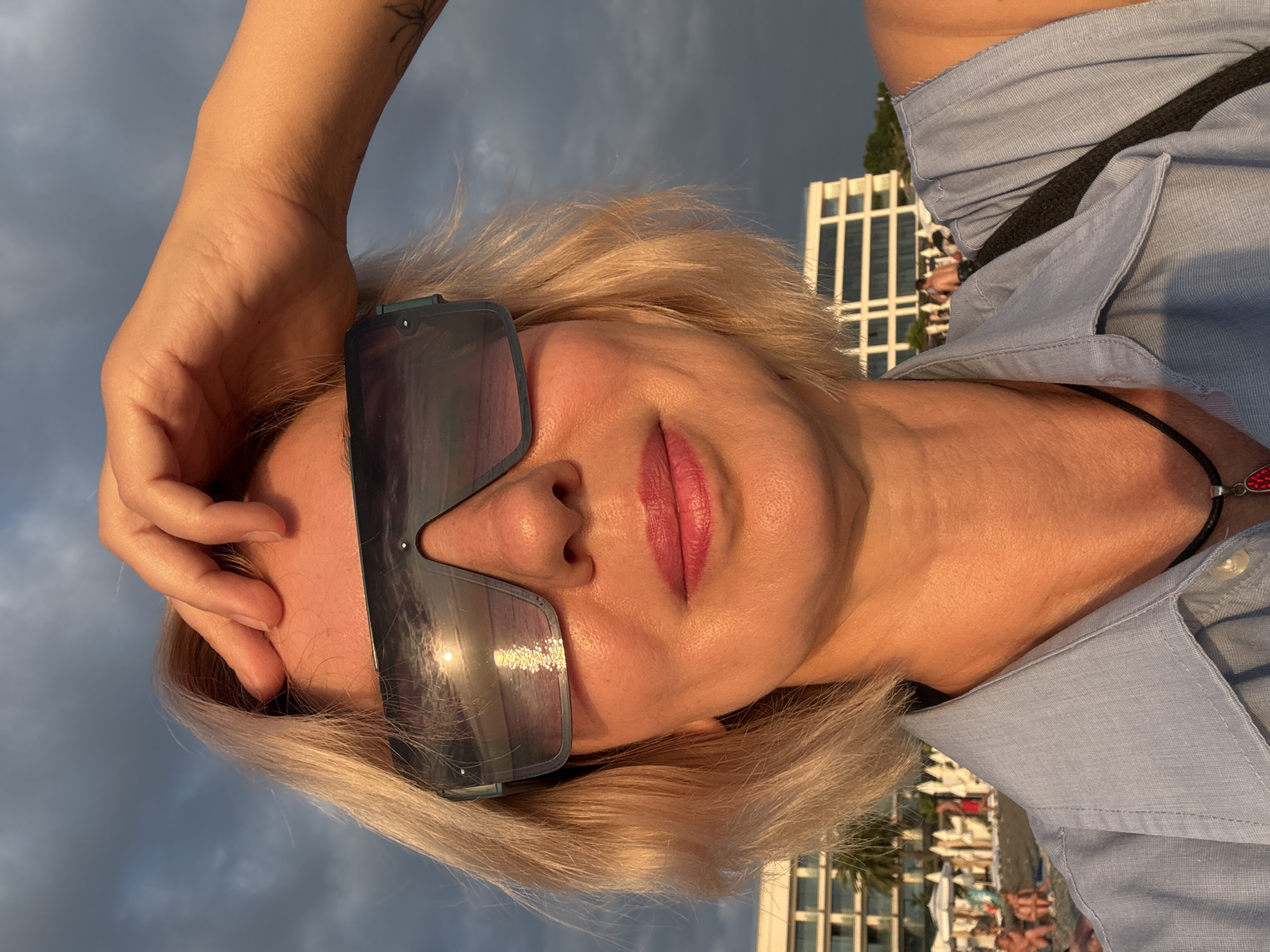 In Sochi, Russia, at sunset, a woman with blonde hair and sunglasses is taking a selfie on a beach. She is wearing a light blue shirt. In the background, there are buildings, palm trees, beach umbrellas, and people enjoying the beach. The sky is cloudy with the sun setting, casting a warm glow on the scene. The reflection of the sun on the water is visible in her sunglasses.
