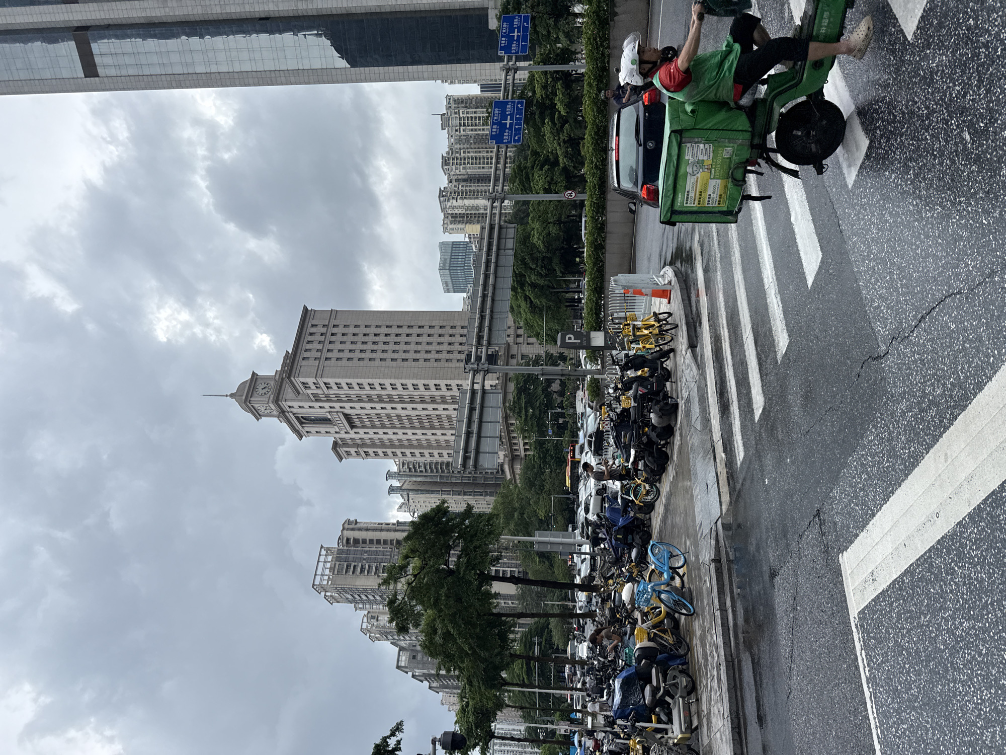 In Tianhe District, China, a delivery person on a green electric scooter, wearing a white helmet and a red shirt with a green vest, rides across a wet pedestrian crossing. The scooter has a large green delivery box on the back. In the background, a variety of parked electric bikes and scooters are lined up on the sidewalk. Beyond the parked vehicles, several modern buildings of varying heights dominate the skyline under a cloudy sky. Traffic signs and trees are also visible.