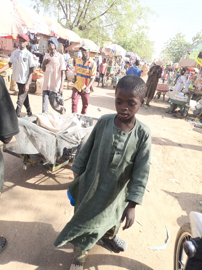 This image depicts a bustling outdoor market scene in Kawaji, Nigeria, under bright daylight.
In the immediate foreground on the right, a young boy with dark skin and short dark hair is standing, looking slightly to his left. He wears a loose-fitting, long-sleeved dark green tunic with buttons and dark sandals. A blue object is partially visible, clutched in his left hand near his hip. To the left of the boy, a portion of a person's foot in sandals and a dark-sleeved arm are visible. A two-wheeled handcart, covered with a light-colored fabric or netting, is situated behind the boy in the mid-ground.
Behind the foreground subjects, a dirt path extends into the background, flanked by numerous market stalls. Three young men are walking from left to right across the mid-ground. The first wears a light polo shirt, dark pants, and a red baseball cap. The second wears a light pink t-shirt, striped pants, and a blue head covering, carrying a dark bag. The third wears a yellow and red patterned t-shirt with "CHRISTIAN DIOR" text, reddish-brown pants, and a light-colored baseball cap.
The background features a dense collection of market stalls with various light-colored awnings, displaying goods that are indistinguishable at this distance. Numerous other individuals are present, walking, standing, and interacting within the market. One person in the distant mid-ground wears a wide-brimmed hat and a long brown garment. Another person further back is pushing a green wheelbarrow laden with items. The ground is unpaved dirt, casting distinct shadows from the bright sunlight. Sparse trees are visible above the market stalls in the far background. A partial view of a white motorcycle wheel is visible in the bottom right corner of the frame.