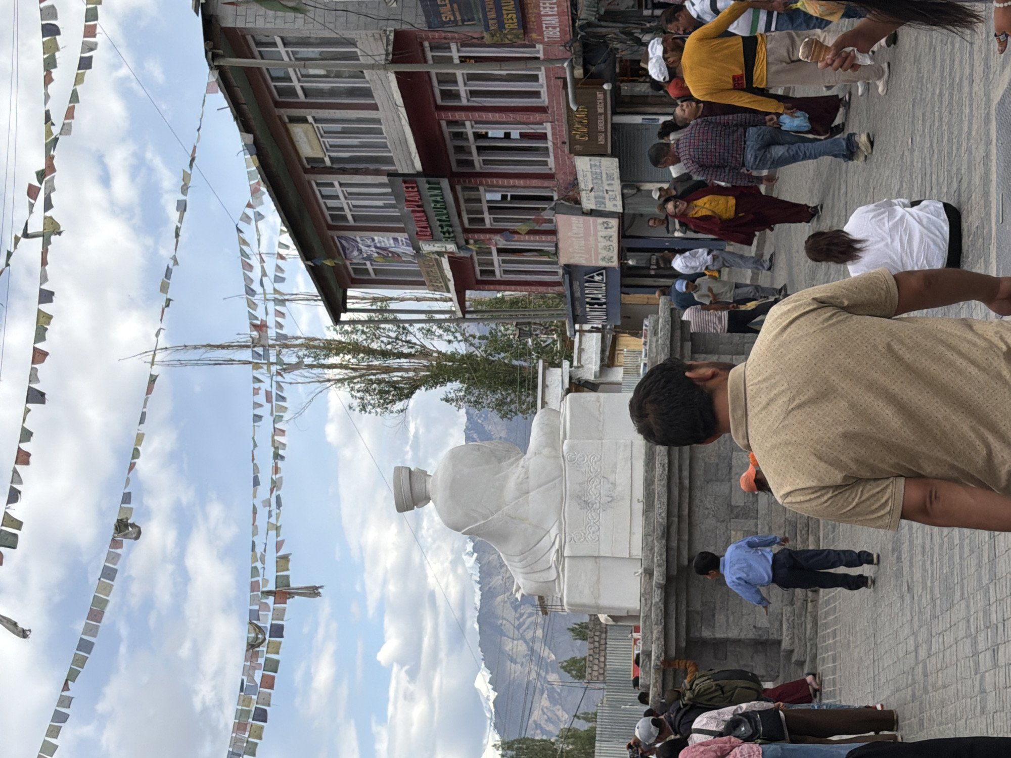 The image depicts a street scene in Leh, India, under a cloudy sky adorned with colorful prayer flags strung across wires. In the foreground, a person wearing a tan, patterned t-shirt is seen from behind, facing away from the camera. To their left, other individuals are visible, some walking and some standing. Dominating the mid-ground is a large, white marble statue of a seated figure, possibly a Buddha, resting on a tiered pedestal. Behind the statue, a two-story building with red-painted walls and white-framed windows stands, featuring various signs, including "NAZWAN PLANET RESTAURANT" and "TIBETAN REFU". Further back, snow-capped mountains form the backdrop. The ground is paved with cobblestones. The atmosphere suggests a busy tourist or public area.