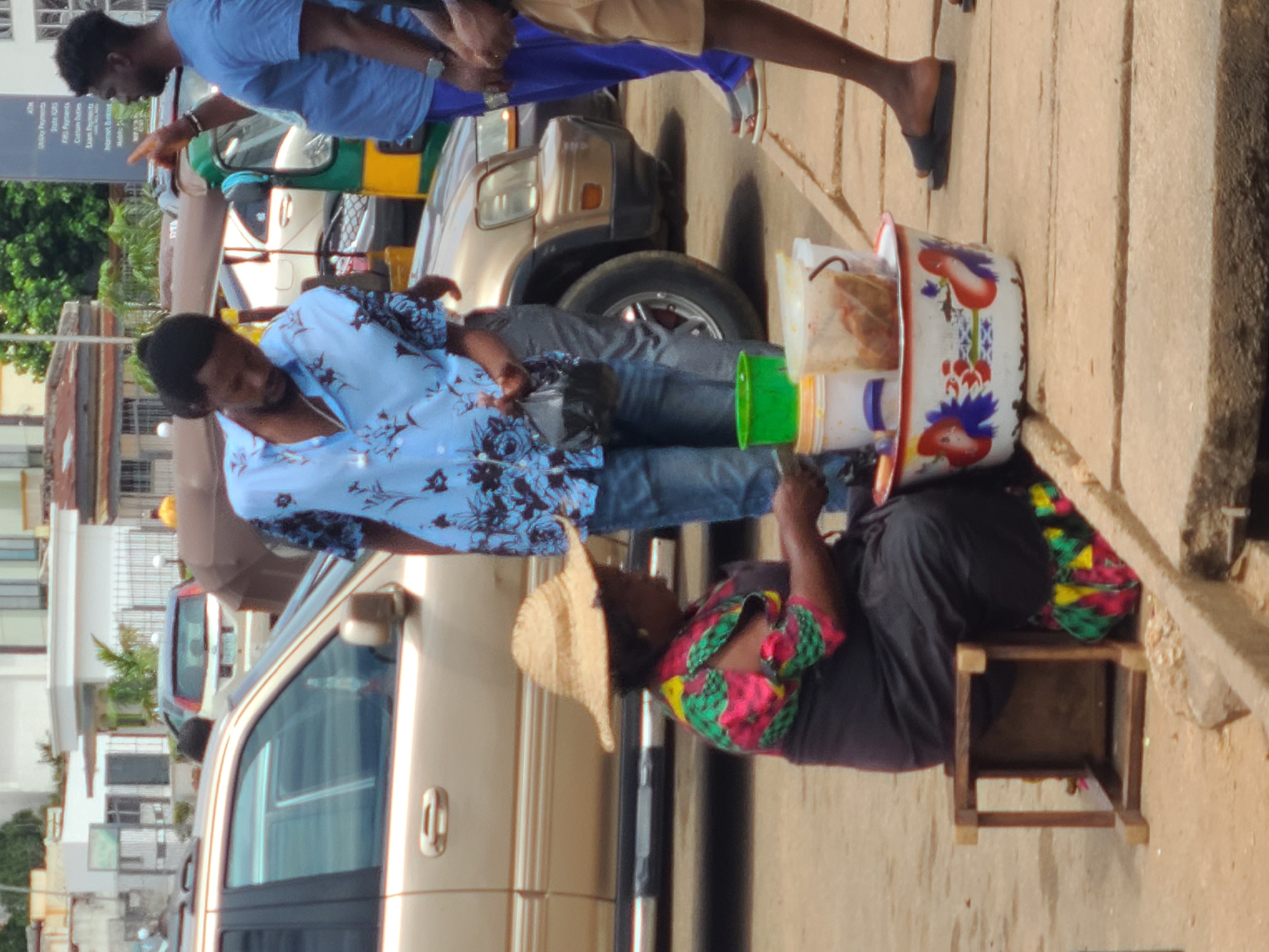An urban street scene in Unknown, Unknown depicts three individuals and multiple vehicles. In the immediate foreground, a dark-skinned woman with a light straw hat is seated on a small, dark wooden stool on a dirt patch, facing right. She wears a brightly patterned short-sleeved top with red, green, and yellow floral designs over a dark skirt or wrap. Before her, on the ground, are multiple plastic containers, including a green bucket and white tubs, and a larger white enamel bowl adorned with red, blue, and green floral patterns. The containers appear to hold various food items.
Directly in front of the woman, a dark-skinned man stands, facing downwards towards the containers. He wears a light blue button-up shirt with dark blue floral patterns, light blue jeans, and holds a black plastic bag in his left hand. To his right, partially cropped, another dark-skinned man stands, wearing a plain blue t-shirt and blue shorts, with dark sandals. He is gesturing with his right hand, pointing upwards and to the right.
Behind the woman and the man in the floral shirt is the front passenger side of a gold or champagne-colored SUV, parked on what appears to be an unpaved shoulder or street edge. Its front wheel, fender, and part of the windshield are visible. In the background, further down the street, other vehicles are present, including a white SUV and a green and yellow three-wheeled vehicle, possibly an auto-rickshaw. Several light-colored buildings with various architectural details line the street. Green foliage and trees are visible above and between the structures. On the far right, a dark vertical sign with white text, listing services like "ATM," "Utility Payments," and "Internet Banking," is partially visible. The scene is illuminated by ambient daylight.