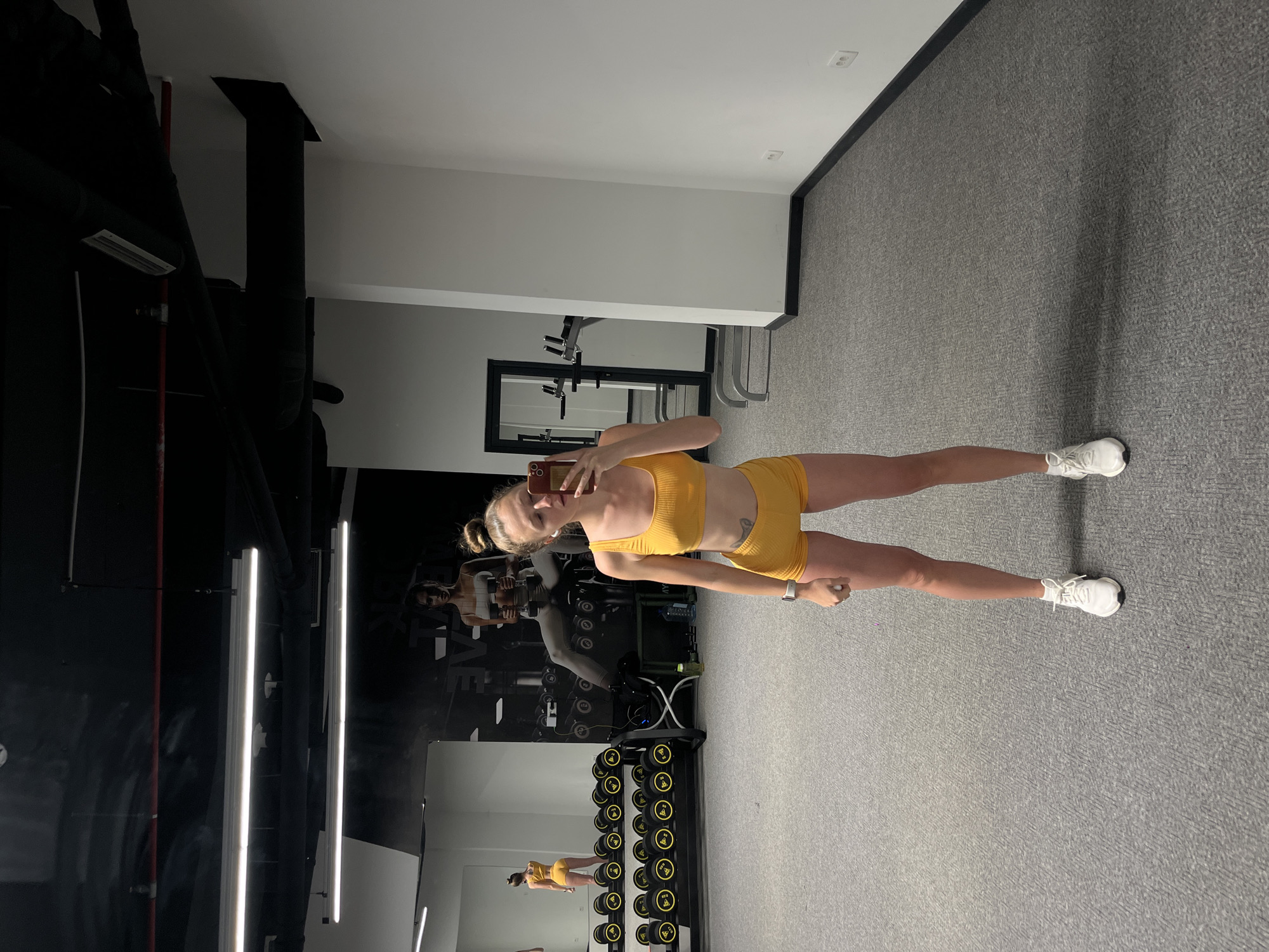 A young woman in a yellow two-piece athletic outfit stands on a gray carpet in a gym, holding a phone to take a selfie. Reflected in a wall mirror are weight racks, workout equipment, and a large wall graphic of an athlete. The gym has white walls and ceilings, accented by black framework. Long, rectangular lights are visible along the ceiling of what appears to be a gym in a modern style. The location is potentially Boreti, Montenegro, based on the user's request.