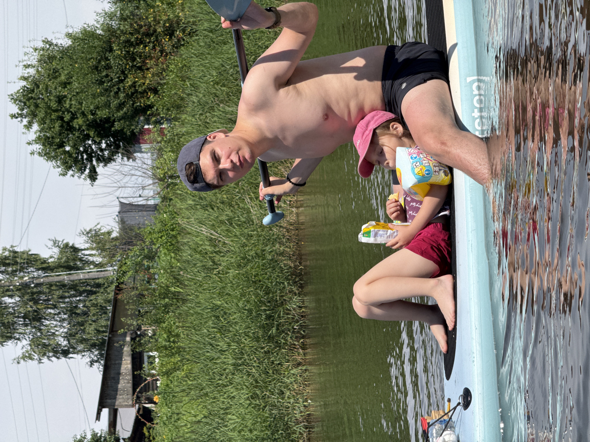 The image shows a young man and a child on a paddleboard in a body of water. The man, wearing a dark baseball cap and black shorts, is holding a paddle. The child, wearing a pink cap and a life vest, is holding a snack. Both are facing towards the right of the frame. The background consists of lush green reeds and trees, with a house visible in the distance. The water is calm, reflecting the surrounding environment. The scene appears to be set on a sunny day. Some text is visible on the side of the paddleboard, which reads "teton". The image is oriented vertically, with the subjects appearing to be moving from bottom to top.