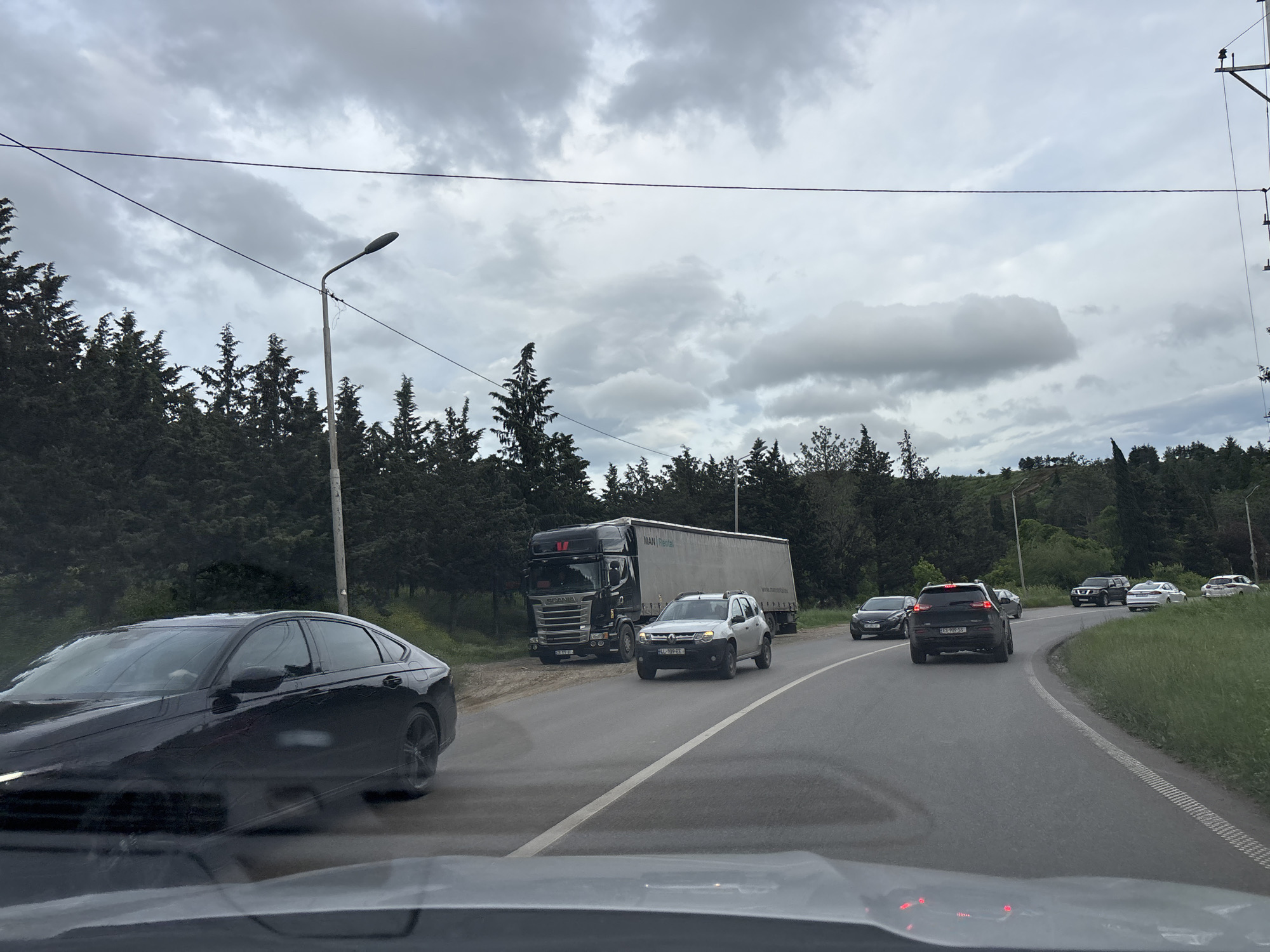The image depicts a traffic scene on a road in the city of Tbilisi, Georgia, under an overcast sky. A black car is partially visible on the left side of the frame. Further ahead, a silver SUV is driving along the road, with a large truck and trailer parked on the shoulder to the left. Several other cars are visible ahead, including a black SUV and white sedan. Trees line the side of the road. A utility pole stands beside the road. The image appears to be taken from inside a vehicle, with the dashboard visible at the bottom of the frame. The license plates of the vehicles are visible. The vehicles are on a road with a white line indicating lanes.