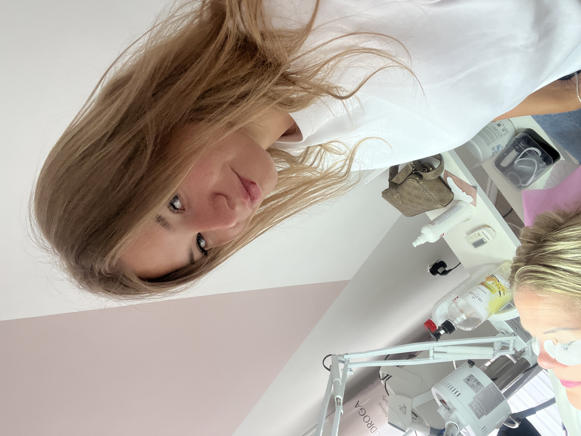 A tilted, indoor eye-level shot features two women. One, with long blonde hair, is looking at the camera, with a neutral expression. She is wearing a white top. The second woman, with blonde hair styled in a bun, is partially visible in the lower right corner, appearing to be working or providing a service, as suggested by the medical instruments and equipment around her. She has white eye protectors on her. The background includes a white wall and furniture, including bottles and equipment likely related to a beauty treatment or medical procedure. The setting appears to be a clinic or salon, with a lamp with the inscription "ODDROGA" and bottles labeled "MULTIPURE" visible. Without further context, it's impossible to confirm the exact location within Budva, Montenegro. However, given the presence of specialized equipment and the professional setting, it's likely a salon or medical spa.