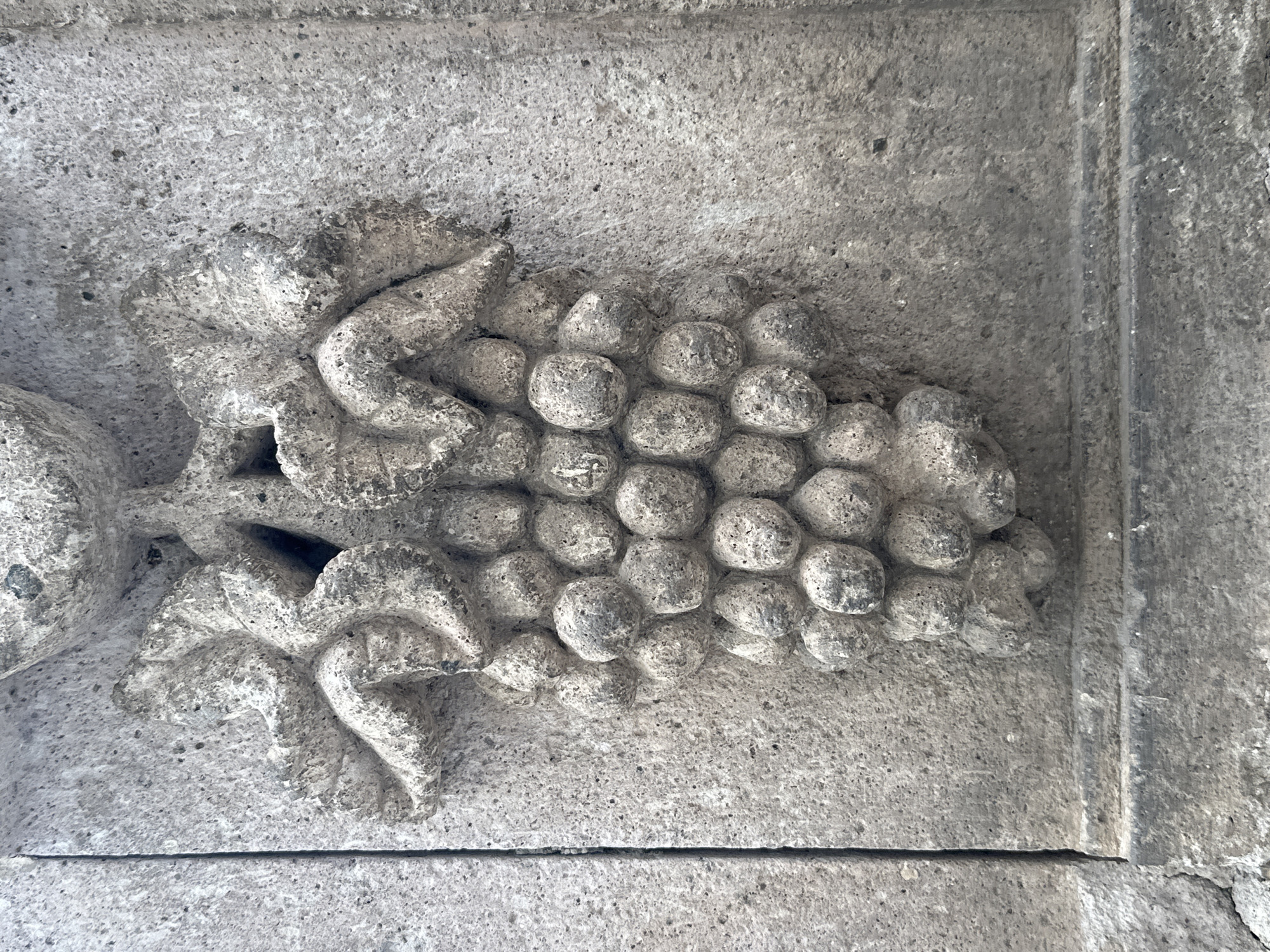 The image is a close-up, eye-level shot of a stone carving depicting a bunch of grapes with leaves, set against a rough, textured stone wall. The carving is detailed, with individual grape shapes and the veins of the leaves visible. The stone appears aged and weathered, with some discoloration and wear. The background wall has a coarse, speckled texture. The lighting is even, highlighting the relief of the carving. This architectural detail is located on the facade of the Museo de la Toma de Zacatecas in Zacatecas City, Mexico.