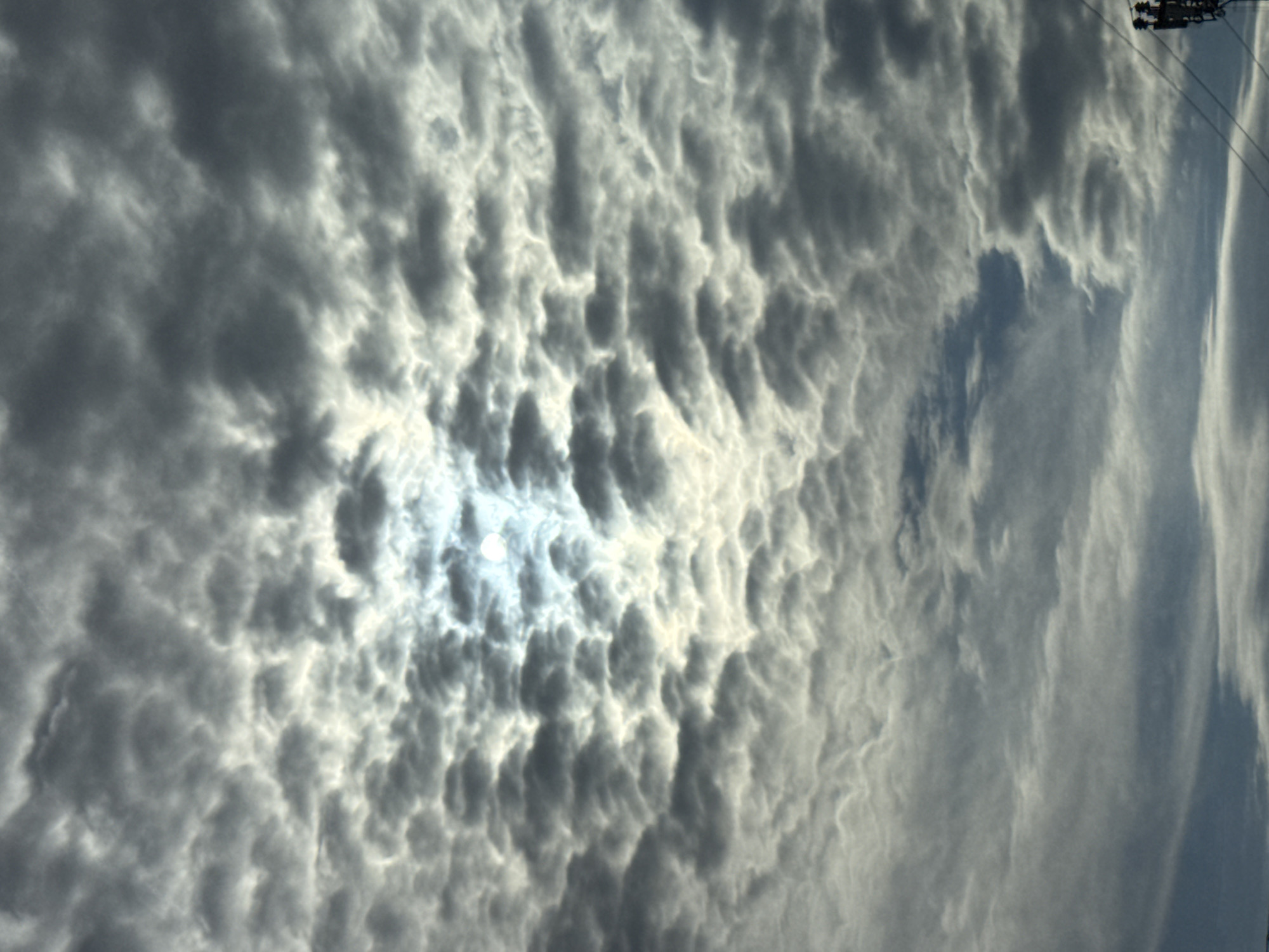 The image displays a dramatic, heavily clouded sky, captured from a ground-level perspective. The scene is dominated by a dense formation of textured, undulating clouds, which appear in various shades of grey, dark grey, and bright white. These formations strongly resemble mammatus clouds or very turbulent stratocumulus, characterized by their distinct, pouch-like or bulbous structures hanging from the cloud base.
A brilliant, diffused light source, likely the sun, is partially visible through a thinner area of the clouds in the upper-left portion of the frame. This intense backlight illuminates the edges of the surrounding clouds, creating bright highlights and emphasizing their three-dimensional, lumpy forms.
In the upper-right corner of the image, elements of human infrastructure are present: a section of a utility pole or power line, with dark equipment and thin wires extending across a lighter patch of sky. This detail confirms the terrestrial setting.
The weather depicted is overcast and atmospheric, suggesting conditions of significant cloud cover, possibly preceding or following a storm. The strong, direct light indicates it is daytime. No people, other specific objects, or activities are visible, and there is no discernible text. The photograph captures a striking natural phenomenon, presumably in Горный, Russia.