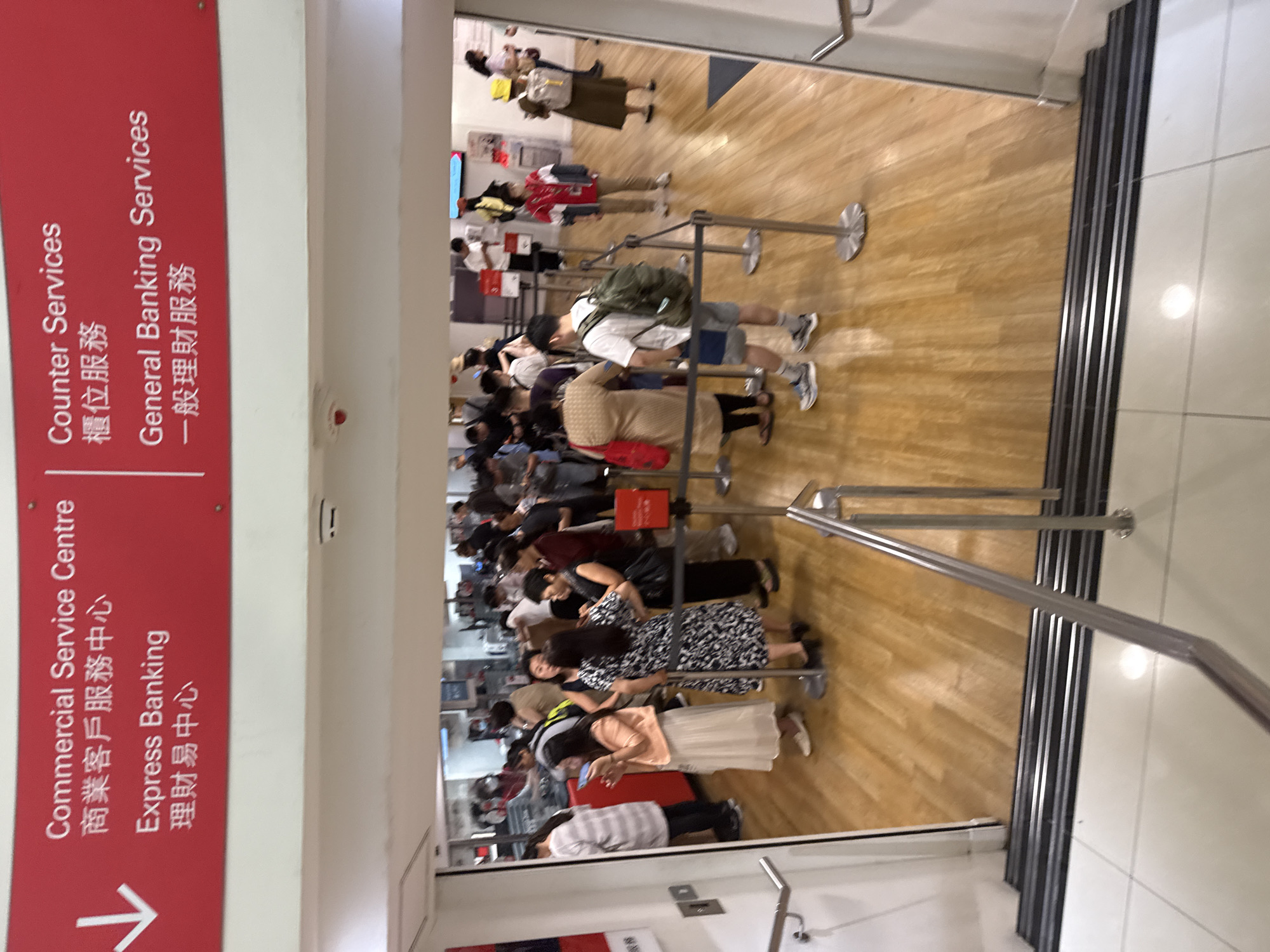 The image shows the entrance to a bank in Hong Kong, China. A red sign with white text indicates various services offered, including "Commercial Service Centre," "Express Banking," "Counter Services," and "General Banking Services." The bank is crowded with people queuing in line. Some individuals are standing, while others are seen talking to each other. The environment appears to be an indoor space with wooden flooring and metal stanchions guiding the queues. The overall scene depicts a busy banking hall.