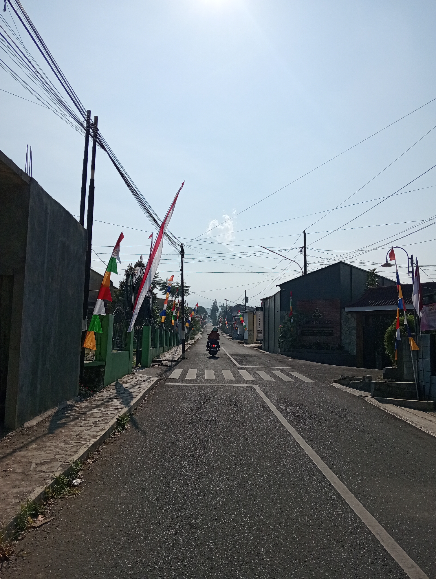 A motorcyclist is riding down a paved road in Pandansari, Indonesia, with the sun in the sky. The road has a crosswalk marked with white stripes. On the left side of the road, there is a gray concrete wall and a green fence adorned with Indonesian flags and banners in red, white, and green. On the right side of the road, there are buildings, some with pitched roofs, and more flags. Overhead, a tangle of power lines crisscrosses the sky. The environment appears to be a residential or commercial street in a tropical or subtropical climate, given the clear sky and vegetation.