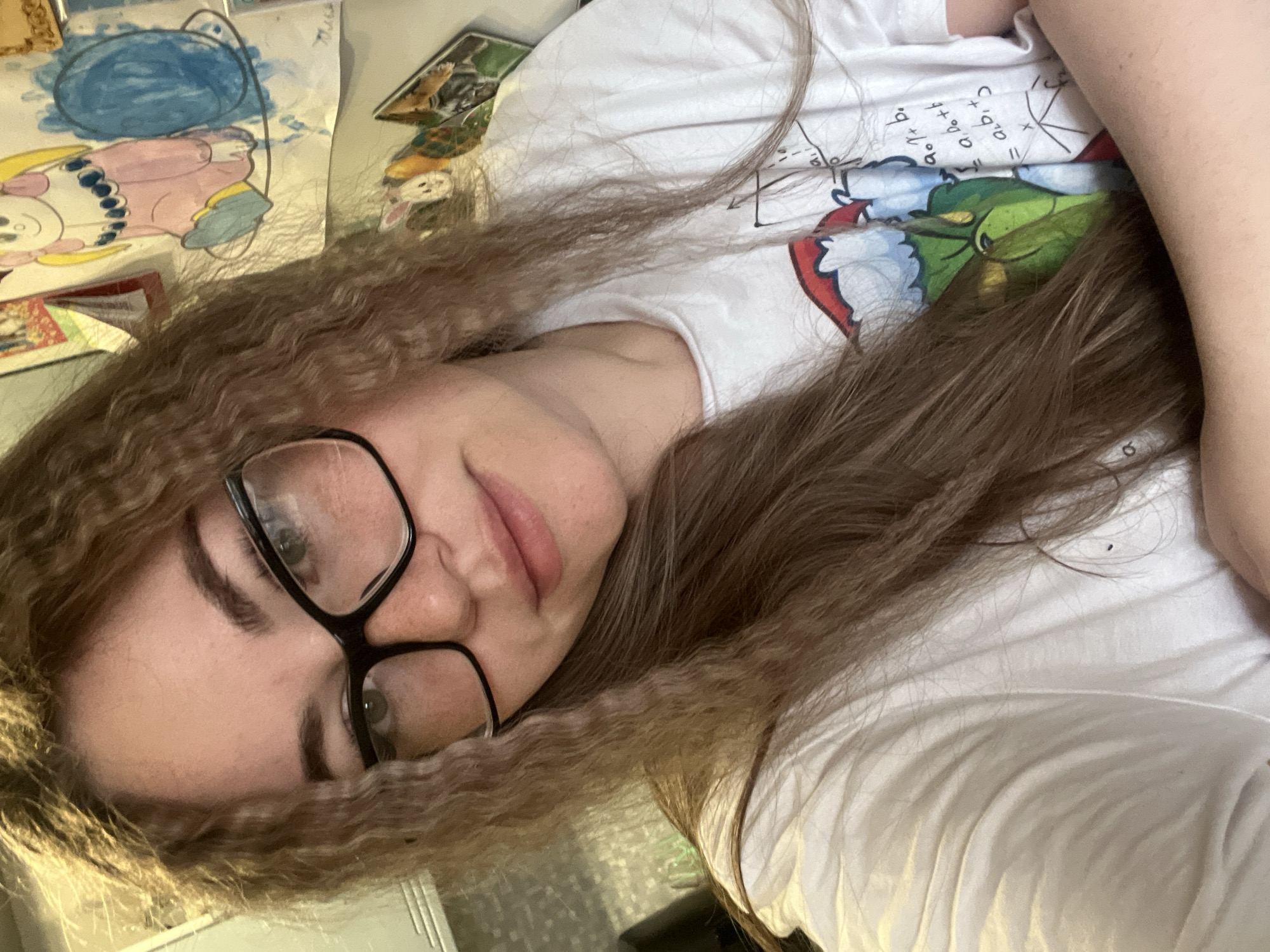 A young woman with long, brown, slightly frizzy hair wearing black-framed glasses and a white T-shirt with a cartoon image is lying down. The T-shirt also has mathematical equations printed on it. There are children's drawings visible in the background. The location in the city of Chaikovsky, Russia, is not discernible from the image.