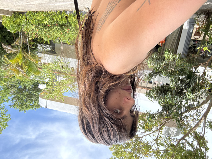 A woman with dark, wet hair and bangs stares to the left with a serious expression. Her skin tone is fair, and a portion of her shoulder is visible. Tattoos are visible on her back and arm. The background features lush green trees and a modern white building with rounded balconies. The sky above is partly cloudy. The overall lighting is bright and sunny.