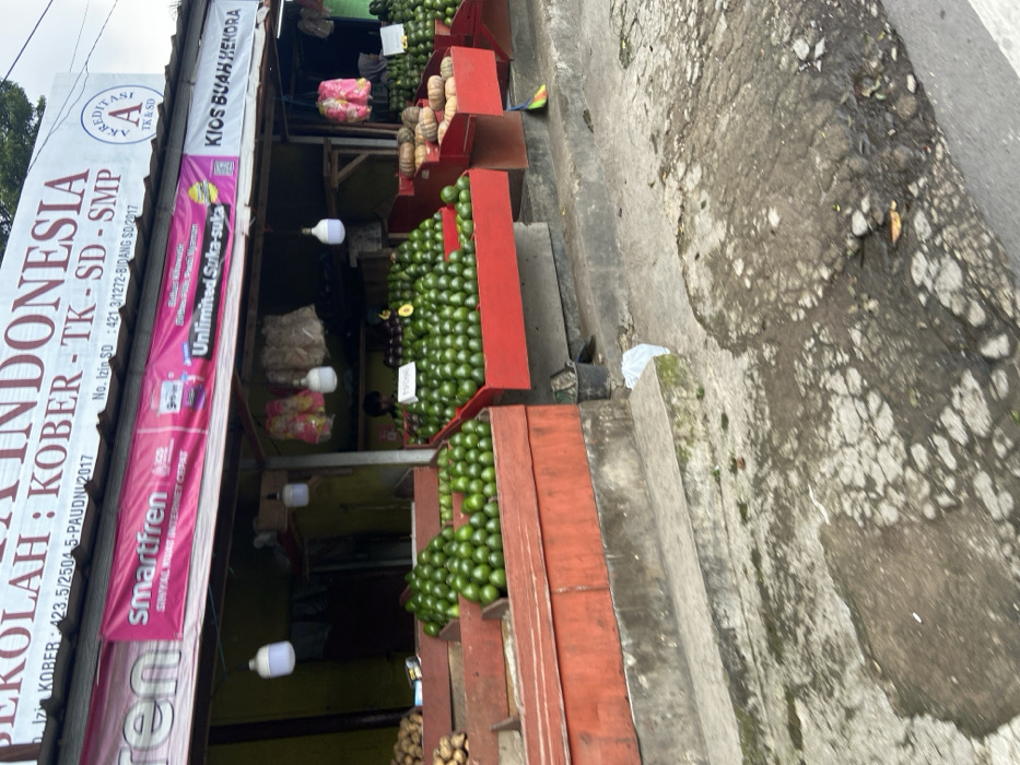 The image shows a fruit stand located in Lembang, Indonesia, at the coordinates -6.818 latitude and 107.613 longitude. The stand is fronted by a cracked, gray concrete sidewalk. The stand is covered by a wooden awning displaying signs in the Indonesian language. The sign on the right states “KIOS BUAH HENDRA.” The sign on the left reads “SEKOLAH INDONESIA KOBBER - TK - SD - SMP” and displays a pink and white “smartfren” logo. The stand is selling green avocados stacked in wooden crates with red borders. Several bags of what appears to be onions are visible inside the stand. There are two white, string lights hanging from the awning above the stand. A person appears to be working inside the stand, but their face is obscured by the counter. A broom is laying on the sidewalk in front of the stand.