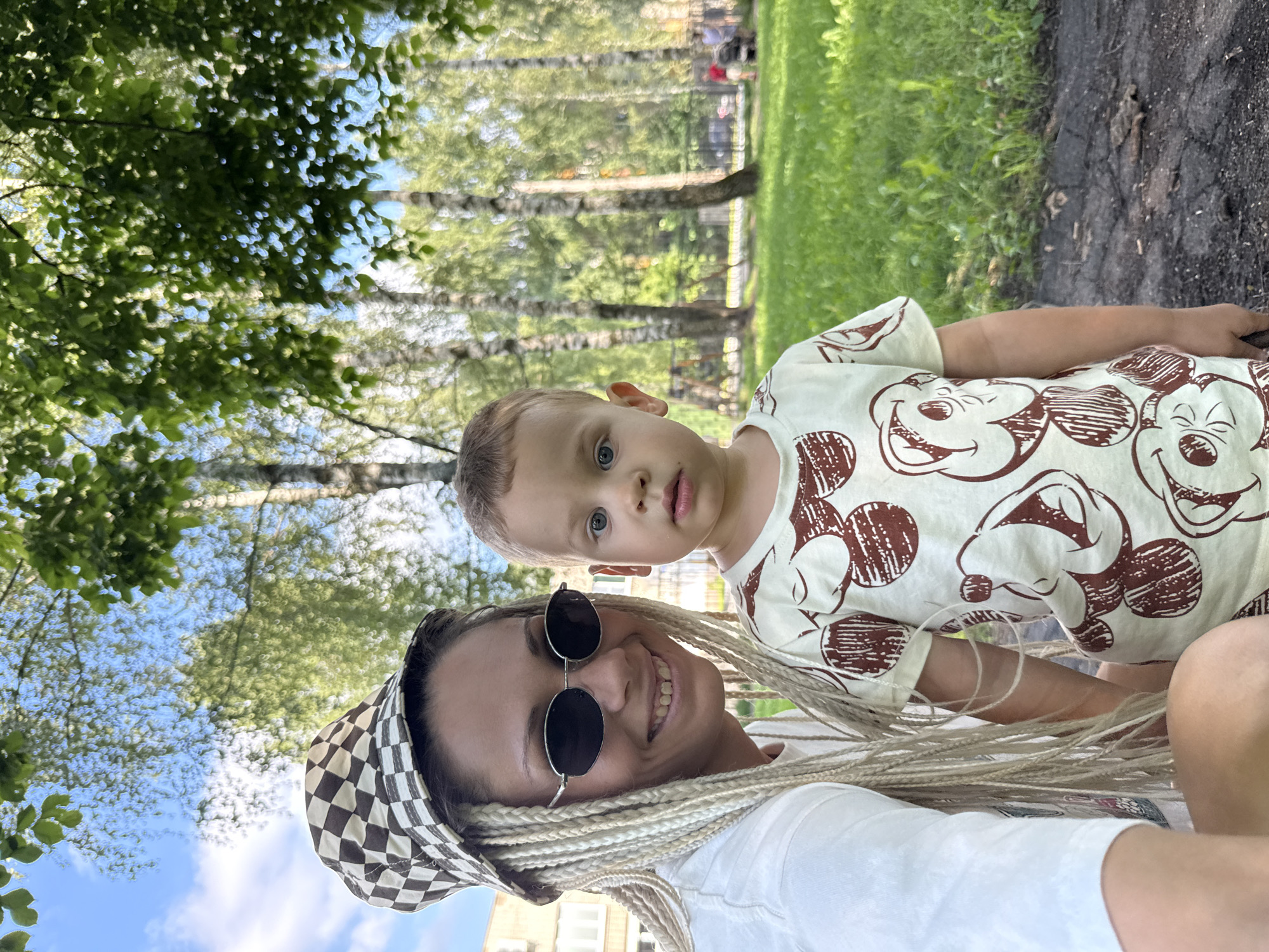 In an outdoor setting, a woman with blonde braided hair and a checkered bucket hat wears sunglasses and a white t-shirt, smiling at the camera. Beside her, a young boy with light brown hair and blue eyes, wearing a t-shirt with a Mickey Mouse pattern, looks directly at the camera. They are positioned in what appears to be a park or recreational area in Rusanovo, Russia, with green grass, trees, and a building visible in the background. The sun is shining, casting dappled light through the leaves of the trees.