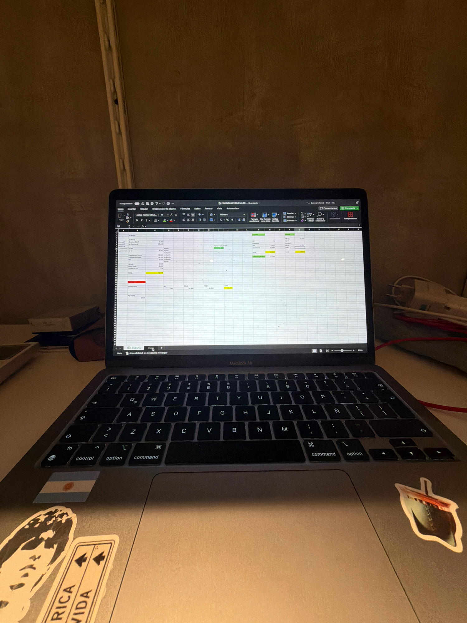 An overhead, eye-level shot shows a closed laptop on a desk. The laptop is a MacBook Air, and its screen displays a spreadsheet program, likely Microsoft Excel. The spreadsheet contains financial data, with columns for different categories and rows listing names and amounts. Various icons and toolbars are visible on the laptop's interface.
The laptop is positioned centrally in the frame, with its keyboard partially visible. The keyboard is dark, with standard QWERTY layout and some Spanish characters, like 'Ñ'. Several stickers are present on the laptop's chassis. On the left palm rest, there's a sticker of the Argentinian flag and another sticker with abstract black and white graphics. On the right palm rest, a sticker depicts a ship.
The background is dimly lit and appears to be a wall with a textured surface, possibly concrete or plaster. The lighting suggests an indoor environment, potentially a home office or study area. The overall scene is devoid of human presence. The image does not contain any identifiable landmarks or features that would place it in North Kuta, Indonesia, or any specific city.