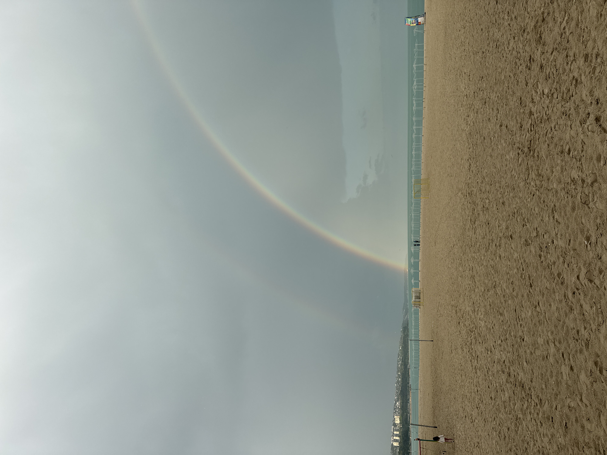 A landscape-oriented photo captures a beach scene under a sky featuring a faint double rainbow. The sandy beach stretches into the distance, occupying the right half of the frame. Small figures are visible on the beach, including a child near the bottom. A line of what appears to be empty sun umbrellas or chairs stands along the border between the sand and the city. The sky, filled with a uniform gray, takes up the left half of the photo. The double rainbow spans the sky, with its vibrant colors contrasting against the dull background. This photo was likely taken in Varna, Bulgaria, based on the beach and architectural styles.