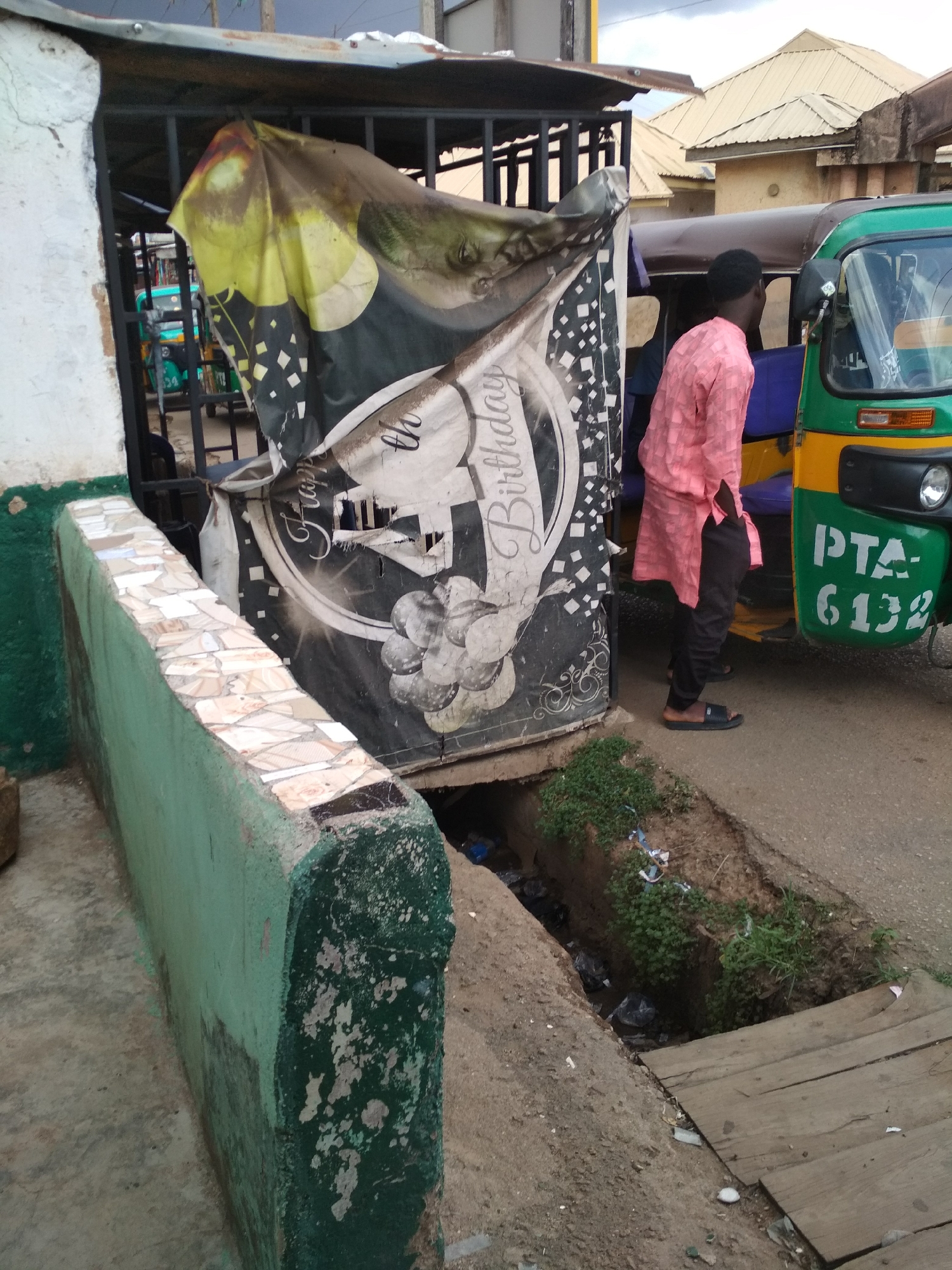 The scene depicts a street in Jos, Nigeria. A three-wheeled motor vehicle, likely a taxi, is parked on the right side of the frame. A man in a pink shirt and dark pants is standing near the vehicle. A sign with a "Happy Birthday" message is partially visible to the left of the man. A small concrete wall with damaged tiling borders a drainage ditch overgrown with weeds and containing trash. A wooden plank covers a section of the ditch.