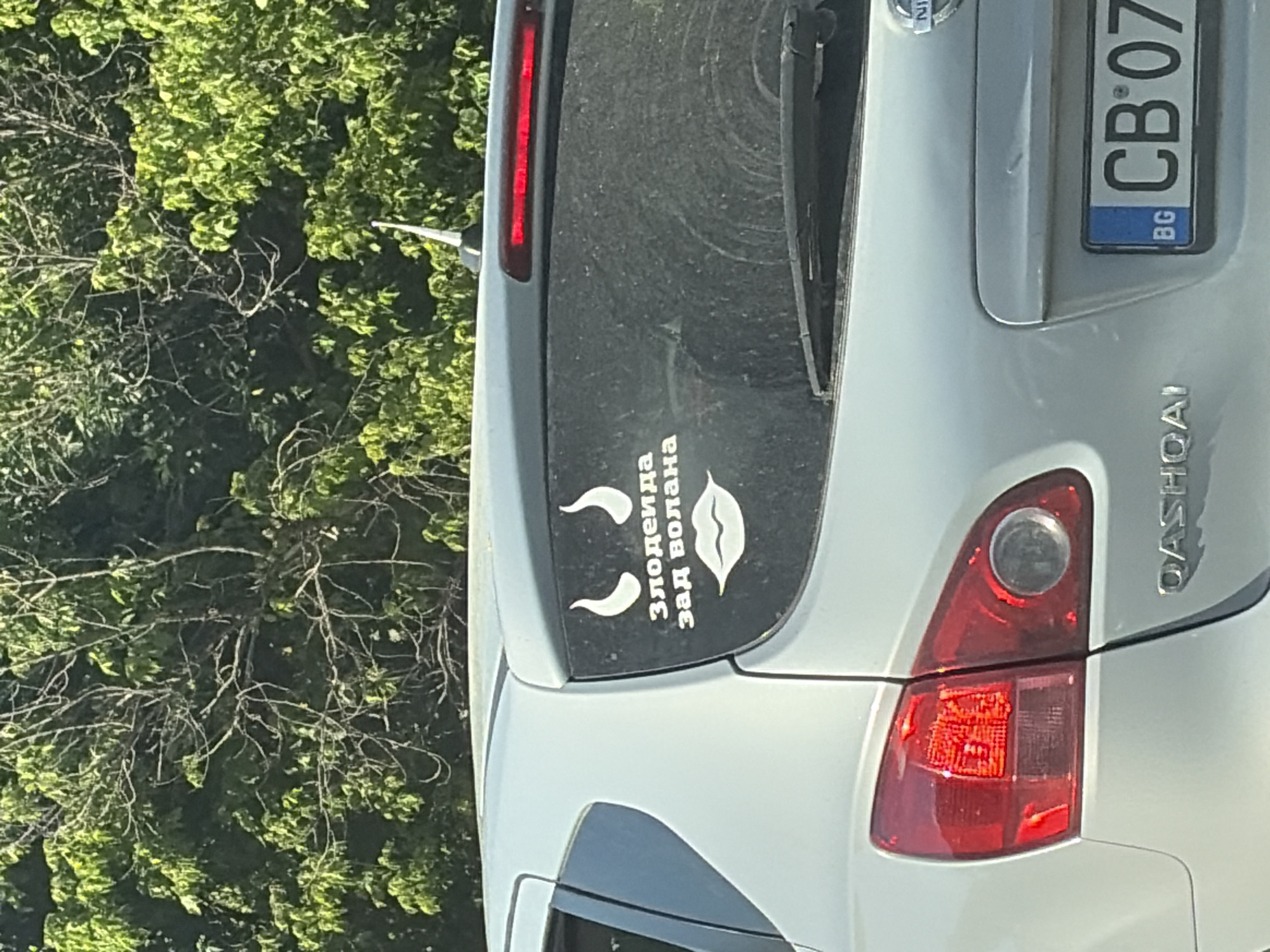 The image shows the rear of a silver Nissan Qashqai car. On the rear window, there is a sticker featuring white decals of devil horns, lips, and text in what appears to be Cyrillic script. The text reads "Злодеида зад волана", which translates to "She-devil behind the wheel." The car has a Bulgarian license plate that reads "BG CB 07". The car is parked outdoors with lush green foliage visible in the background, suggesting a sunny day. The car's taillights are red and clear. The rear windshield wiper is visible. The license plate is positioned centrally on the lower part of the rear bumper. The car is clean, with no visible damage. The overall setting is outdoors in a natural environment, possibly a street or parking lot with trees.