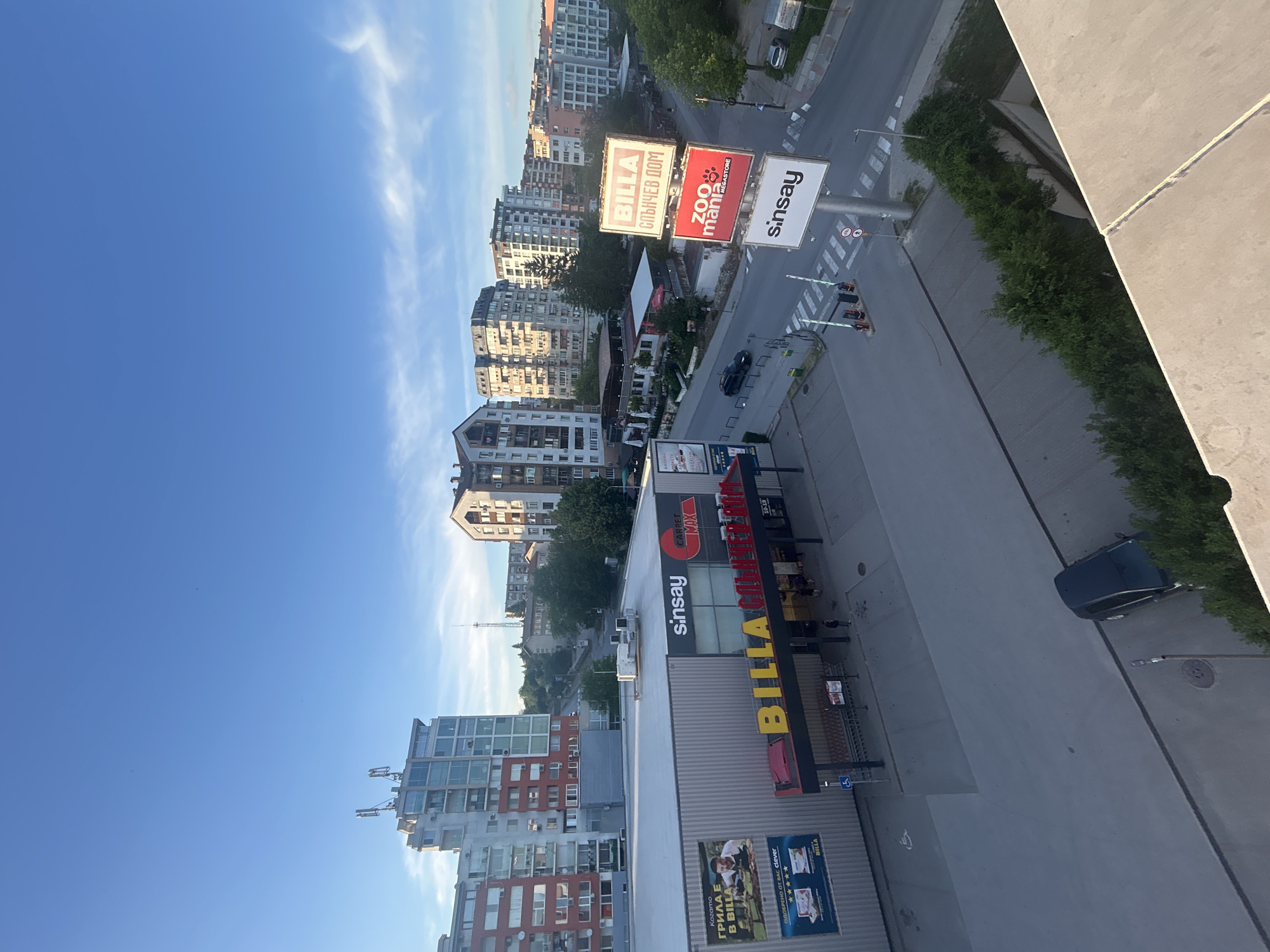 This is an exterior shot of the city of Veliko Tarnovo, Bulgaria. The scene is observed from a higher elevation, capturing a mixed urban landscape under a clear blue sky with scattered clouds. Dominating the foreground are commercial buildings, identifiable by signage of stores. "BILLA", "sinsay", "Zoo Mania MEGASTORE" are visibly prominent on the buildings. The surrounding environment is an urban area with a mix of residential buildings of varying heights and architectural styles. Roads and streets are visible, with limited vehicular traffic. The scene is generally well-lit, and the photo conveys a sense of a calm, yet active urban environment.