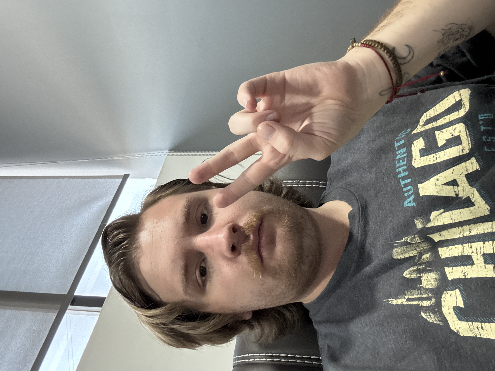 A person is pictured from a low angle, appearing to be in a seated position. They are fair-skinned with light brown hair and a beard/mustache. They are wearing a dark gray t-shirt with yellow and blue text. They are holding up their hand in a peace sign gesture near their face, while another hand is held up in an "OK" sign. Their left arm has a few tattoos and bracelets. The location appears to be indoors, possibly an office space, given the wall and window blinds. There is no information to suggest they are in Cuautitlán Izcalli, Mexico.