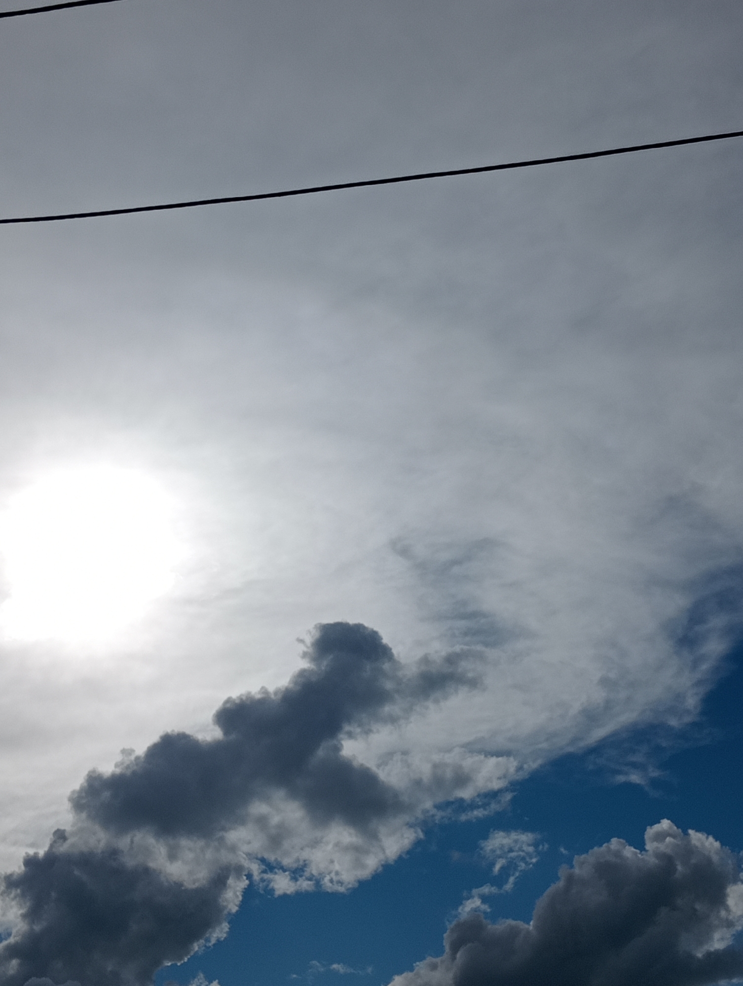 The image shows a sky with clouds and two power lines running horizontally across the top of the frame. The sun is visible on the left side of the image, appearing as a bright, glowing orb obscured by some clouds. There are dark, fluffy clouds in the lower part of the image, contrasting with lighter, wispy clouds and the blue sky. The scene is a sky view with no specific identifiable landmarks or context related to a city.