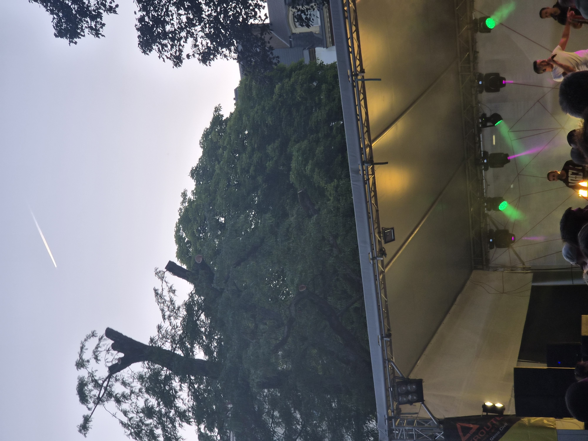 In the twilight, an outdoor stage is set up for a performance. The stage is covered by a white canopy, illuminated from above by yellow lights. Multiple spotlights are mounted on the stage's structure, some emitting green and pink beams into the hazy air. On the stage, a male singer in a white t-shirt stands center, holding a microphone and gesturing with his right hand. To his right, another male musician plays a guitar, and to his left, a third man stands. A crowd of people is gathered in front of the stage, partially visible in silhouette. A large banner hangs on the left side of the stage, featuring a triangular logo and text. Trees and buildings are visible behind the stage, under a pale sky where a contrail from an aircraft is present. The scene is taking place outdoors in Differdange, Luxembourg.