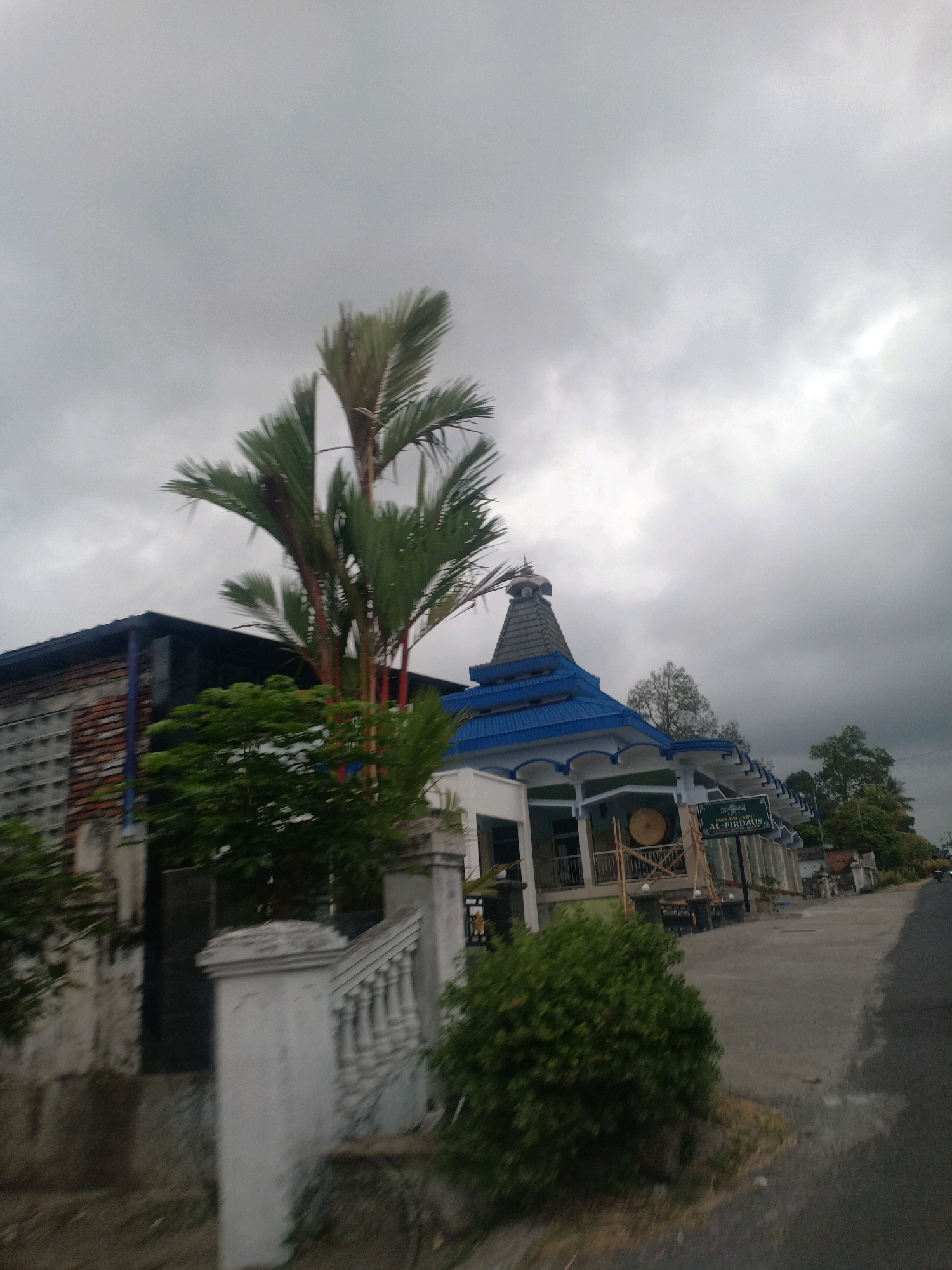This image, taken in Kloposawit, Indonesia, depicts the exterior of a mosque and its surrounding environment under an overcast sky. The mosque, identified by signage as "MASJID JAMI' AL-FIRDAUS," features a distinctive blue roof with white trim and a conical dome topped with a finial. The building has large windows and an open-air veranda. In front of the mosque, a large, circular drum, likely a kentongan, is visible, supported by scaffolding.
To the left of the mosque, a building with a brick and glass block facade is partially visible. Several trees and dense green foliage are present in the foreground and midground. A white stone balustrade and pillar are prominent in the lower left quadrant of the image, suggesting a pathway or entrance. The scene is captured from a roadside perspective, with a paved road extending into the distance to the right, bordered by vegetation and other structures. The overall impression is one of a humid, possibly tropical, atmosphere due to the cloudy sky and lush greenery. There are no people visible in the frame.