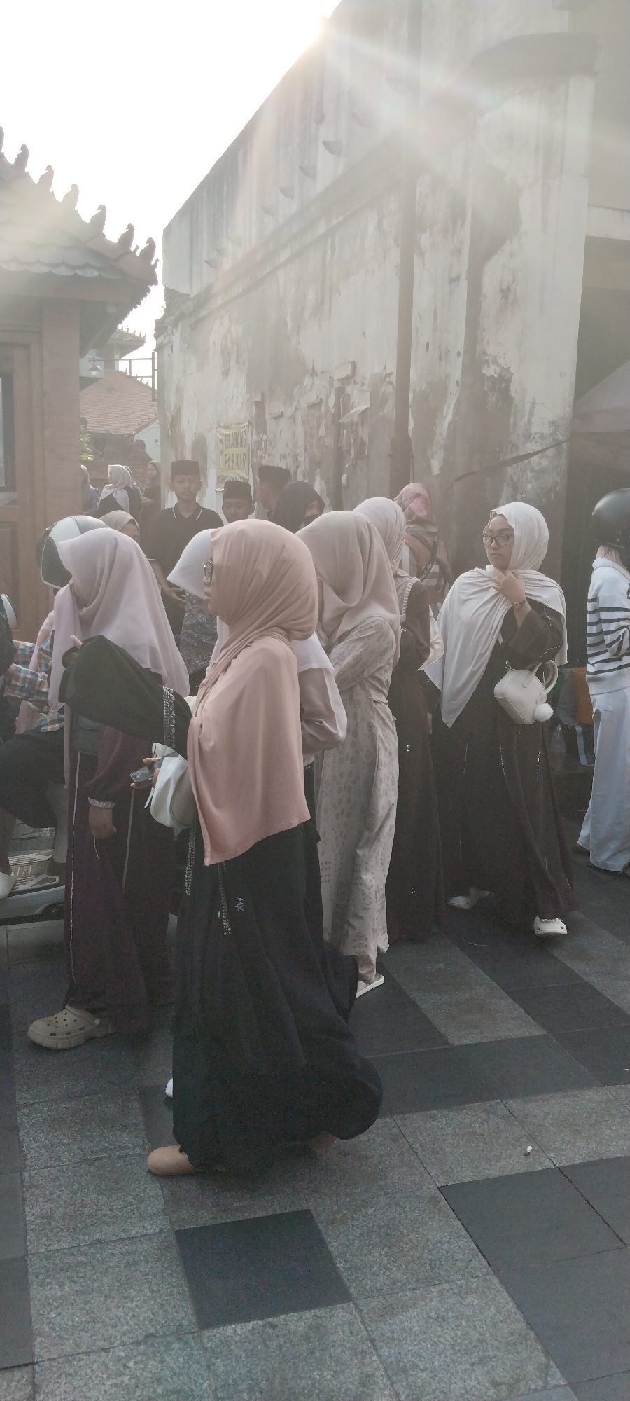 This is a street-level photograph taken in the city of Kauman, Indonesia, during daylight hours, possibly in the late afternoon due to the strong backlight and lens flare. The scene depicts a gathering of people, many of whom are wearing hijabs and modest clothing, suggesting a culturally conservative environment. The architecture visible includes traditional Indonesian tiled roofs and a weathered, textured wall. The ground is paved with patterned tiles. The overall impression is that of a public space where people are socializing or waiting.