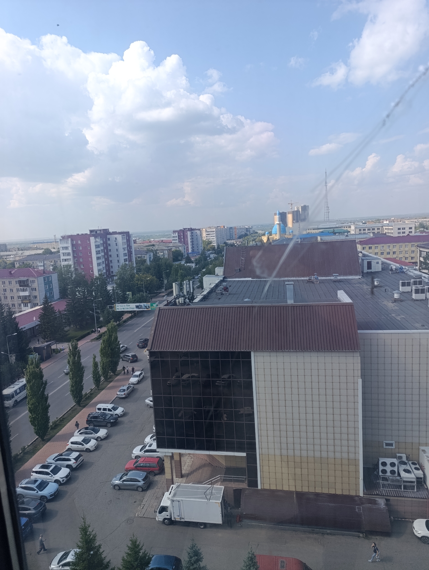 This aerial view captures a street scene in Petropavl, Kazakhstan, on a partly cloudy day. The foreground features a large building with a brown sloped roof and a beige tiled facade, with large reflective windows. In front of this building, a white truck is parked, along with several other vehicles including a white car, a red car, and a silver SUV. Further back, a parking lot is filled with numerous cars of various makes and models, predominantly sedans and SUVs. A street runs parallel to the parking lot, with traffic including a white bus and several cars. Pedestrians are visible on the sidewalks and near the vehicles. Behind the immediate buildings, a cityscape unfolds with residential buildings of varying heights and architectural styles, some painted in purple and white, and others in lighter colors. A large radio tower stands in the distance, alongside construction cranes. The sky is a blend of blue and white clouds, with a faint aerial wire or cable crossing the upper portion of the image. Green trees line the street and are interspersed throughout the urban landscape.