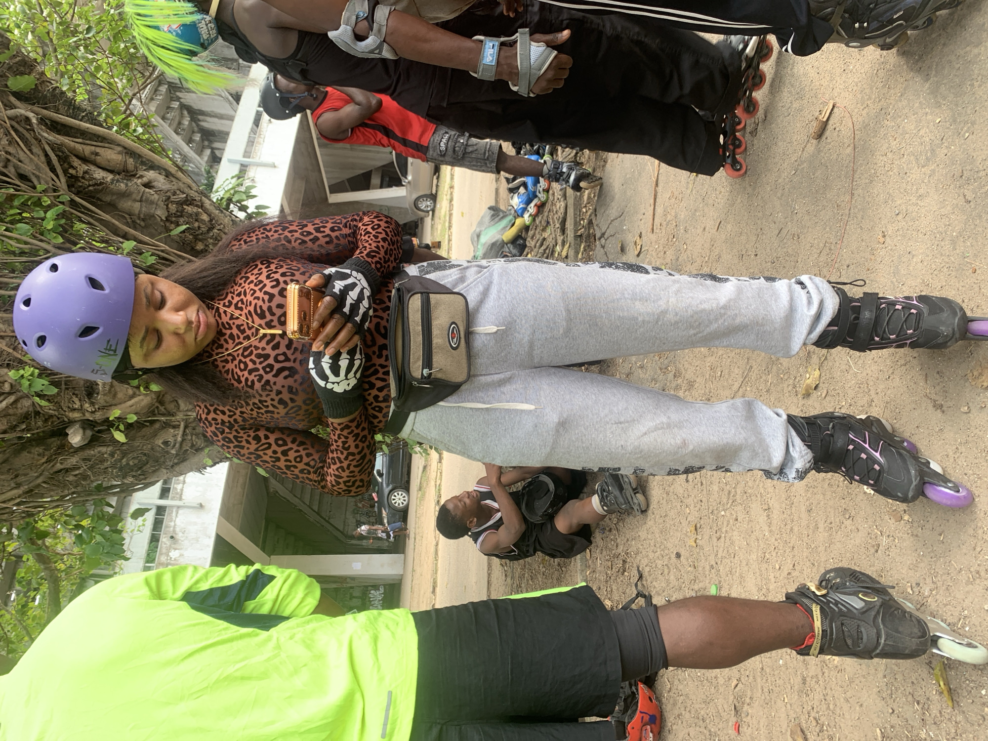 In Alagbede, Nigeria, a group of individuals are gathered outdoors, possibly near a road or pathway, as suggested by the presence of a vehicle in the background and the dusty ground. A young woman wearing a purple helmet and a leopard-print top is the central figure, focused on her phone. She has on black and white fingerless gloves and grey sweatpants with a fanny pack around her waist. She is also wearing rollerblades. To her left, a person in a bright neon-yellow jacket and black shorts stands, also wearing rollerblades. To her right, another person in a black tank top and black pants is visible, with protective gear on their arms and forearms, and wearing rollerblades. In the middle ground, a young person in a black tank top and shorts is sitting on the ground, holding a helmet. In the background, more people and rollerblades are scattered, indicating a group activity. The environment appears to be outdoors, with trees and some structures visible in the distance, including what looks like a bridge or overpass.