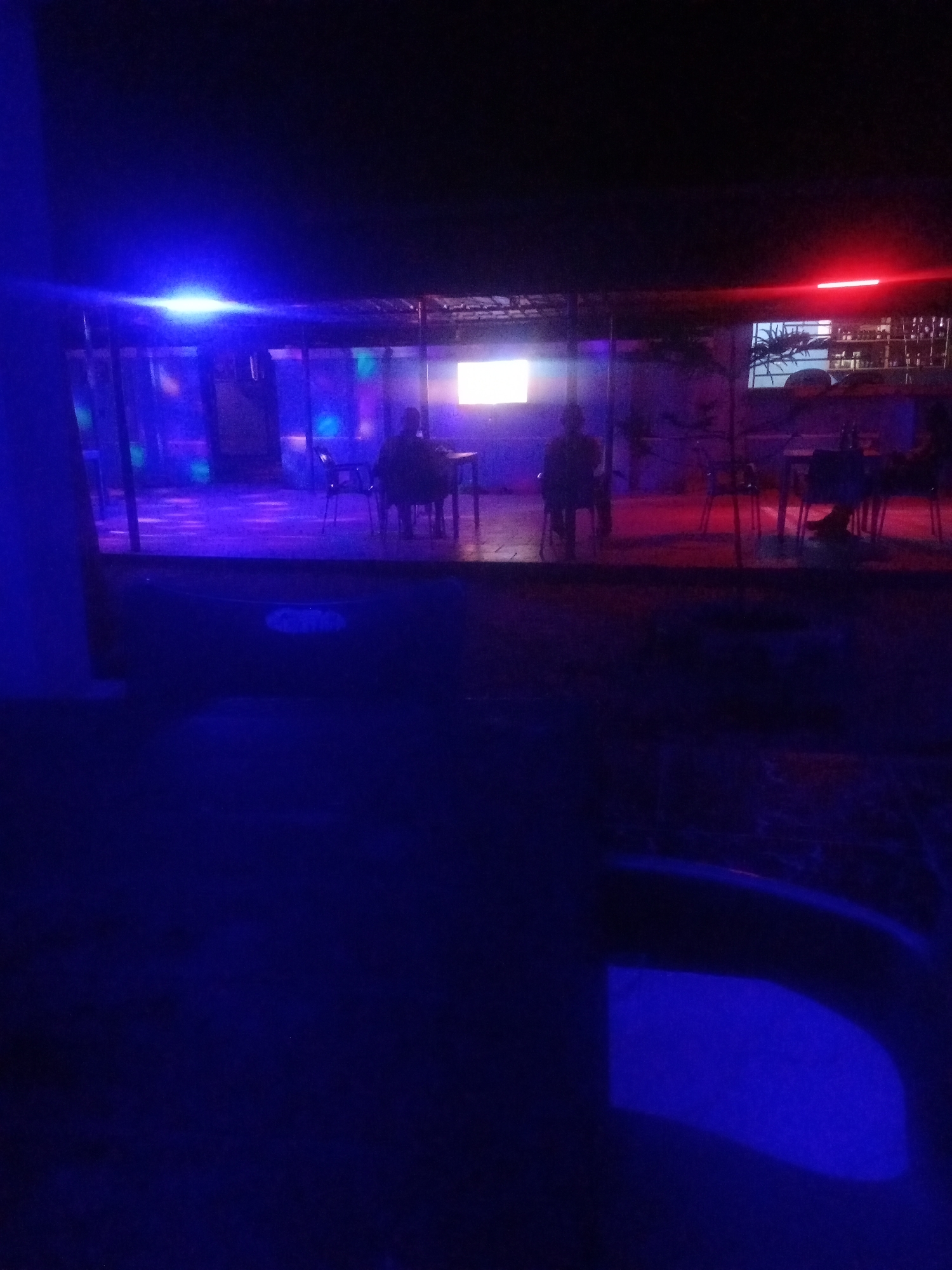 The image depicts an outdoor seating area at night, illuminated by blue and red neon lights. In the background, a covered structure with glass walls reveals a room with tables and chairs, where two individuals are seated. One person is visible from the back, facing a television screen. The environment appears to be a dimly lit establishment, likely a bar or restaurant, in Sabon Gari, Nassarawa, Nigeria. The foreground is dominated by a dark, unlit area with a reflective blue surface, possibly water or a wet floor.