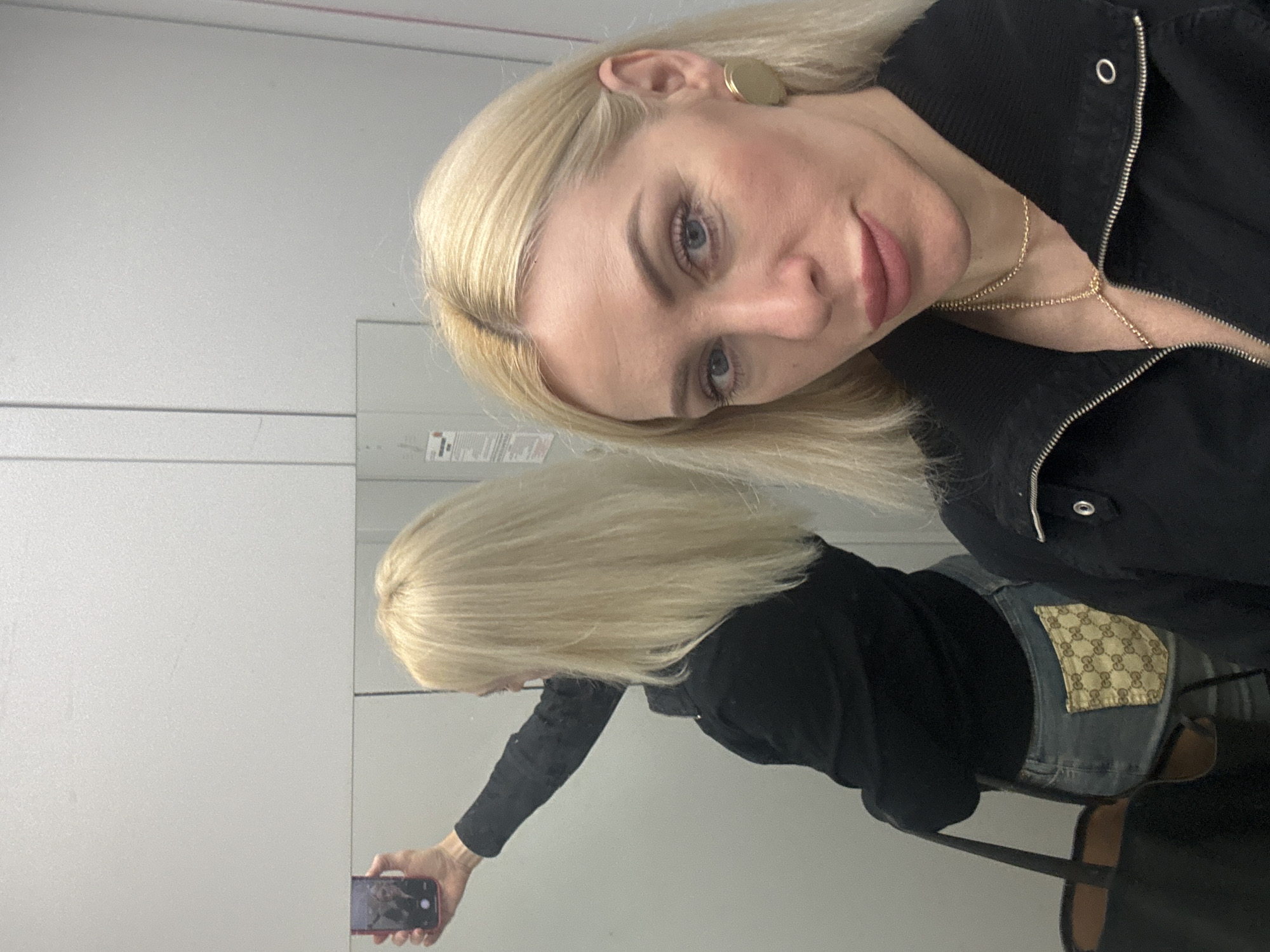 A woman with blonde hair is taking a selfie in a mirrored room. She is wearing a black jacket and a gold necklace. In the mirror, she is wearing denim pants with a patterned Gucci logo patch. She is holding a phone in her hand to take the photo. The environment is a room with white walls.