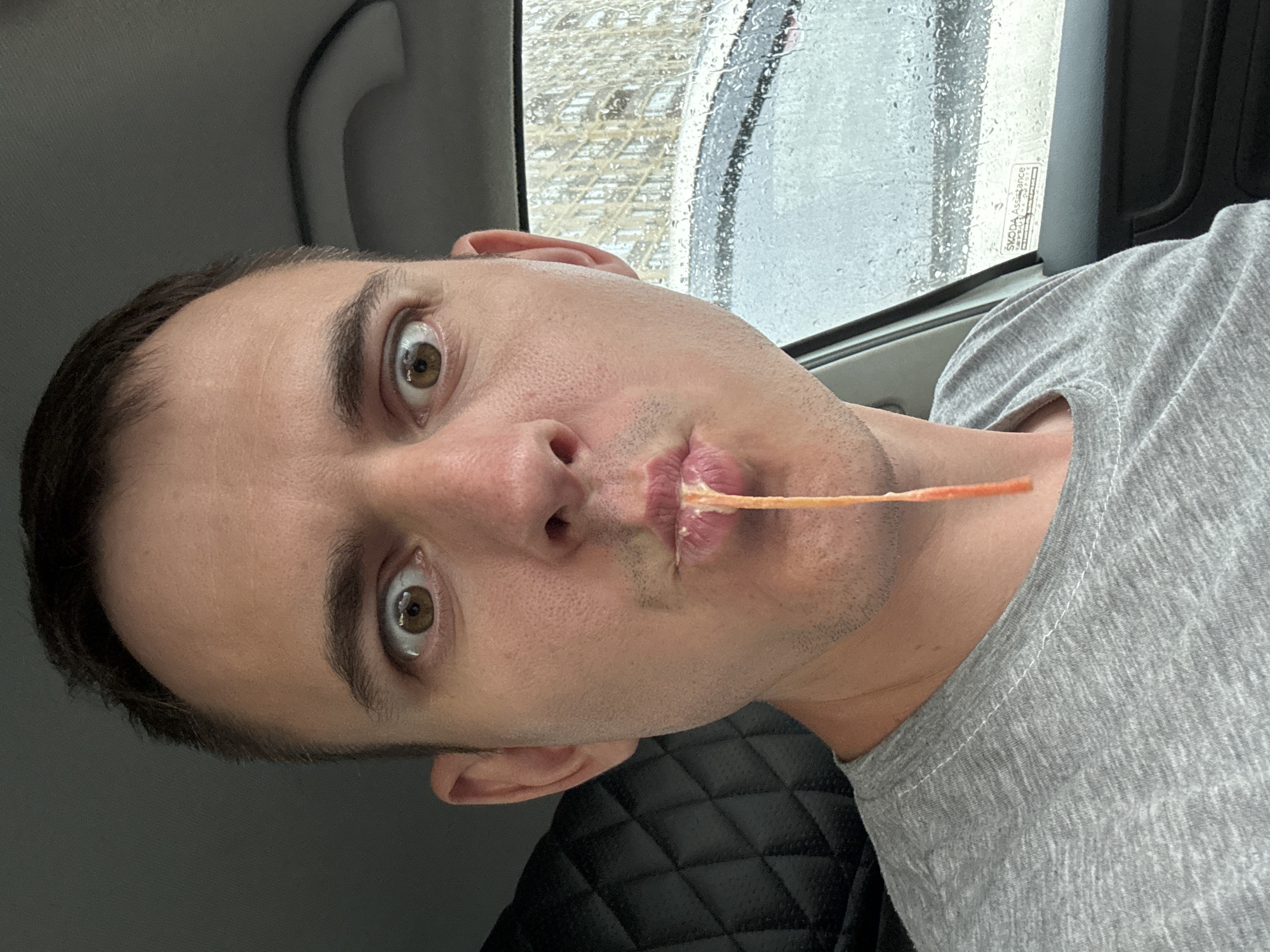 A selfie of a man with brown eyes and dark hair, wearing a gray t-shirt, taken inside a car. He has what appears to be a cheese puff in his mouth. Outside the car window, it is raining, and a building with a latticework facade is visible. A sticker on the window reads "ŠKODA Assistance." The seat appears to be made of black quilted leather. Based on the sticker information and visual analysis, the photo was taken in the city of Krasnodar, Russia.