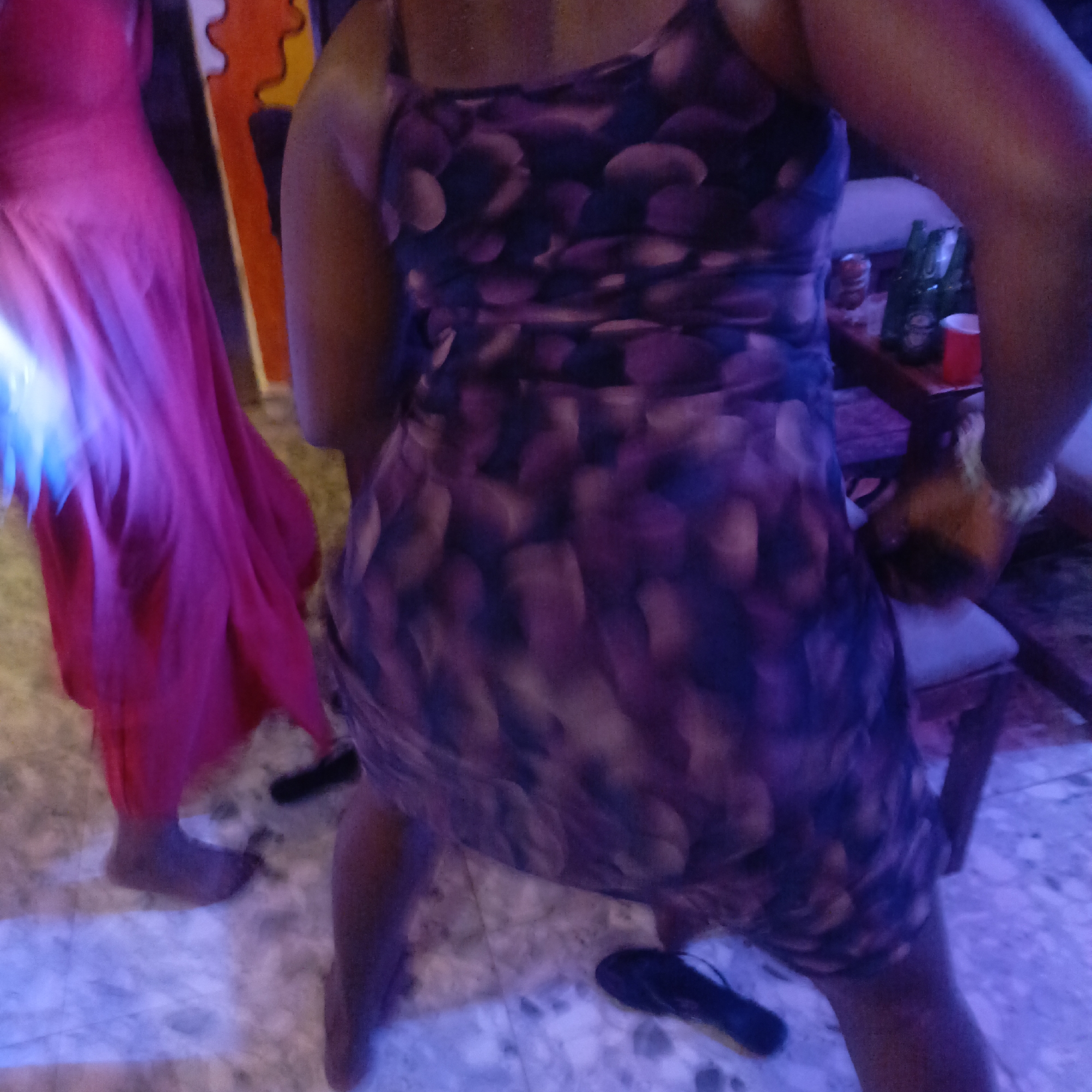 The image shows two people indoors, likely in a private residence in Akwanga, Nigeria. One person is wearing a pink dress, while the other is wearing a purple and blue patterned dress. The person in the patterned dress is closer to the camera. There's a small table behind the person in the patterned dress, with what appear to be beer bottles and a red cup on it. A chair is also visible next to the table. A pair of black flip-flops are on the floor near the person in the patterned dress. The lighting appears to be a mix of indoor lighting and possibly a colored light source. The floor is tiled. An abstract wall hanging is partially visible in the background.