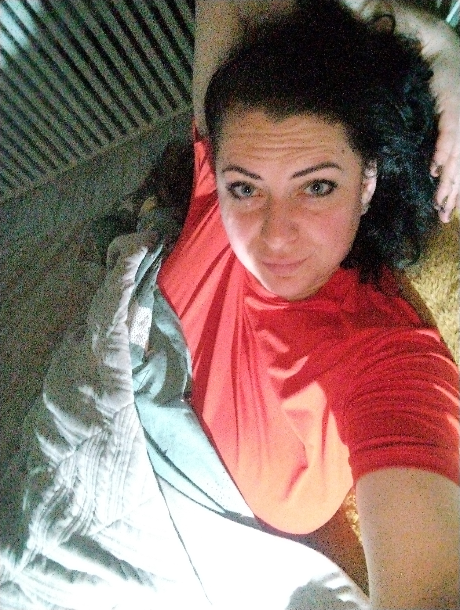 A woman with dark curly hair and blue eyes is lying down wearing a red shirt. She appears to be on a bed or floor covered with a blanket, and there is a textured surface near her head, possibly a carpet or rug. Behind her, in the upper left, there is a slatted or barred structure. There is no information to establish the location of this scene in the city of Grishanki, Russia.