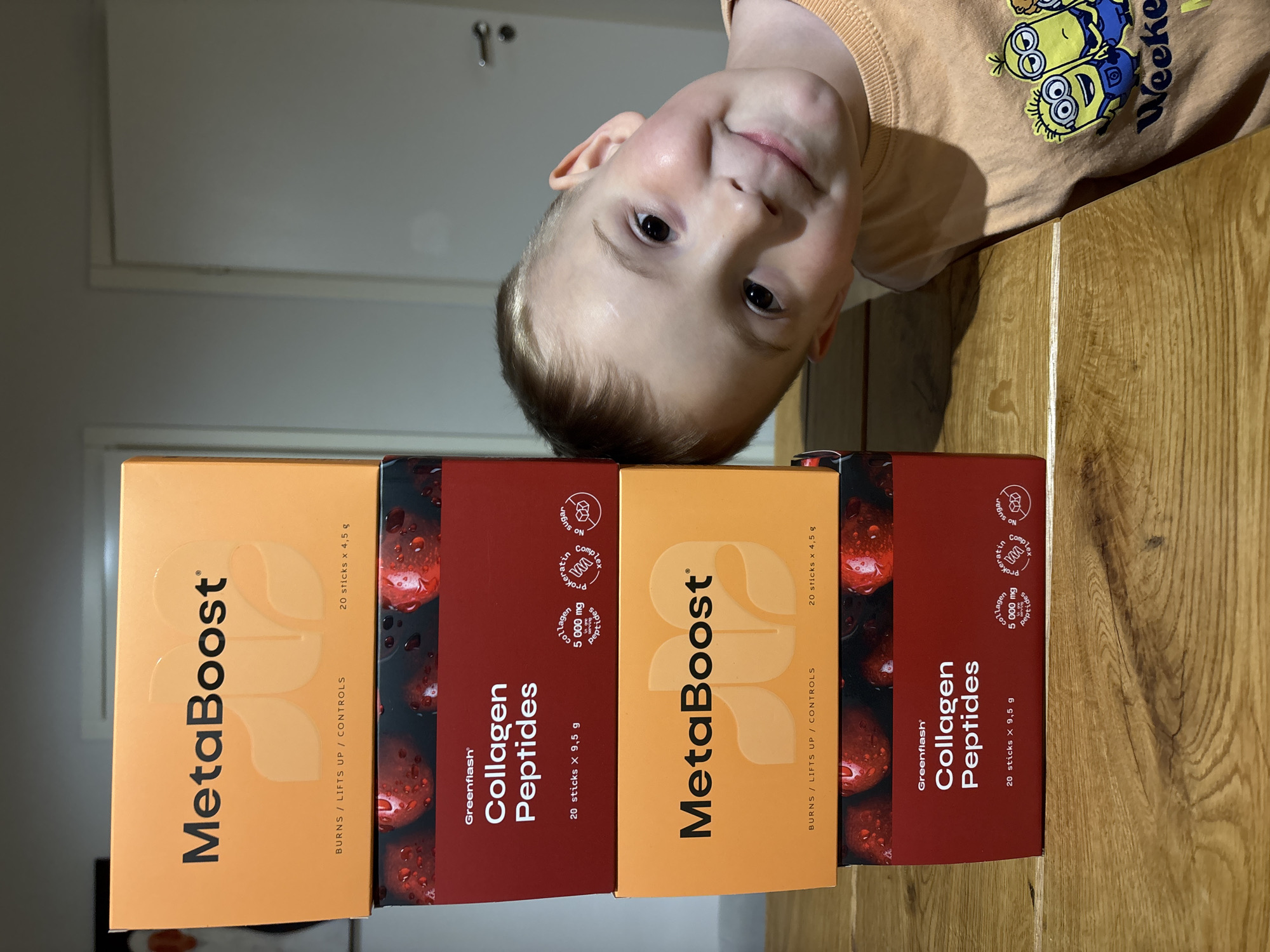 A child with fair skin and short blonde hair is looking downwards toward the camera. He is wearing a light orange t-shirt with a Minions design. Below him, stacked on a wooden table, are four boxes of Greenflash supplements. From left to right, there is a box of MetaBoost, a box of Collagen Peptides, another box of MetaBoost, and another box of Collagen Peptides. The boxes of MetaBoost are orange, and the boxes of Collagen Peptides are red. The background is out of focus, but it appears to be a room with white walls and a light-colored door. There is no indication of location in the city of Chaikovsky, Russia.