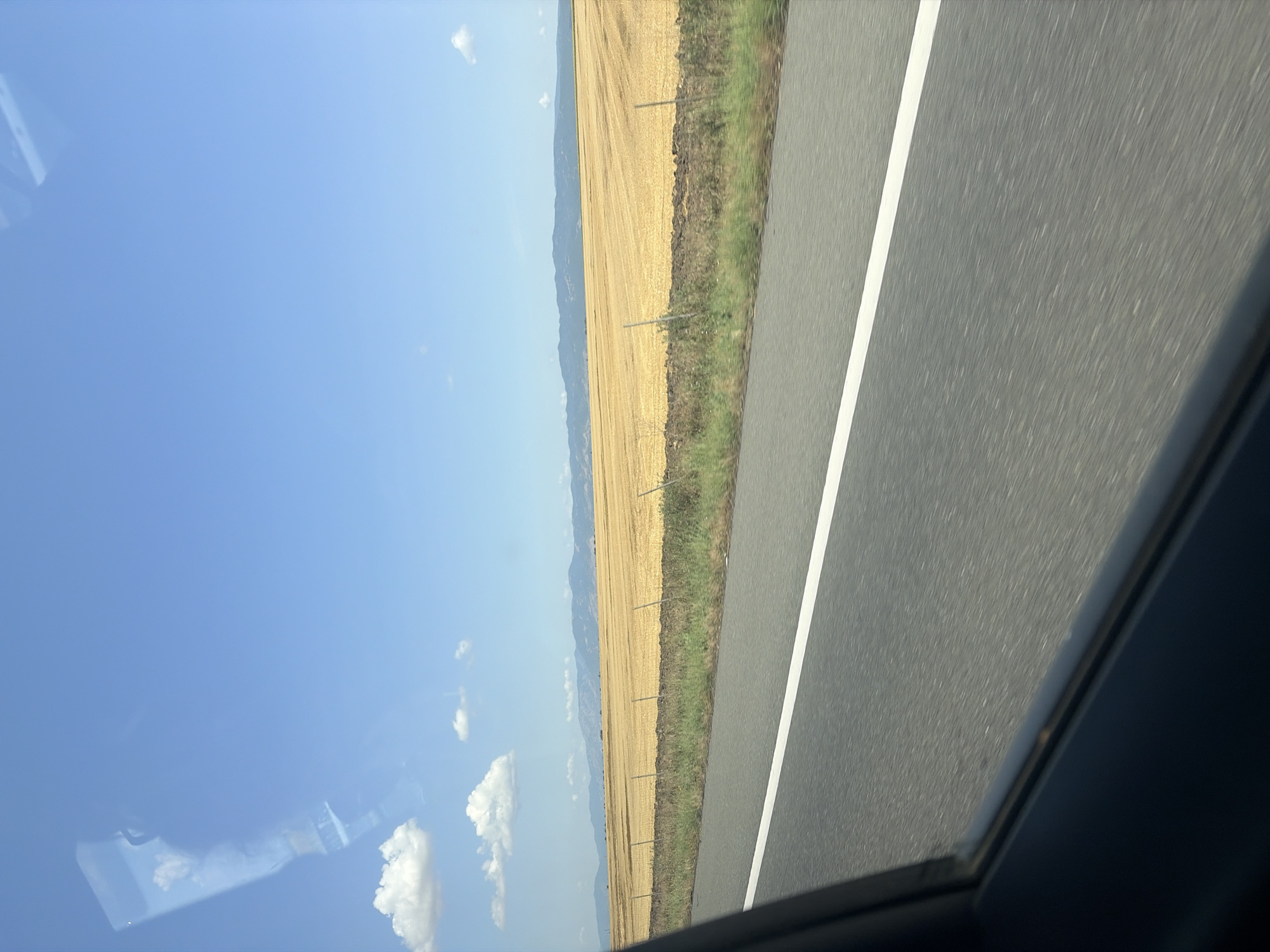 The image depicts a landscape scene, likely taken from a moving vehicle, as indicated by the blurred motion of the road in the foreground and the view through a car window. The scene features a vast, golden-colored field, possibly harvested wheat or barley, stretching towards a range of distant, hazy mountains under a clear blue sky with scattered white clouds. A dark line of vegetation and a simple fence run along the edge of the field. The overall impression is one of rural tranquility and open space. There is a reflection of what appears to be a bottle and clouds in the upper left quadrant of the sky, likely from the car's window. The image is not specific to the city of Nedyalsko, Bulgaria, or any other identifiable urban location, and no people or specific actions are visible.