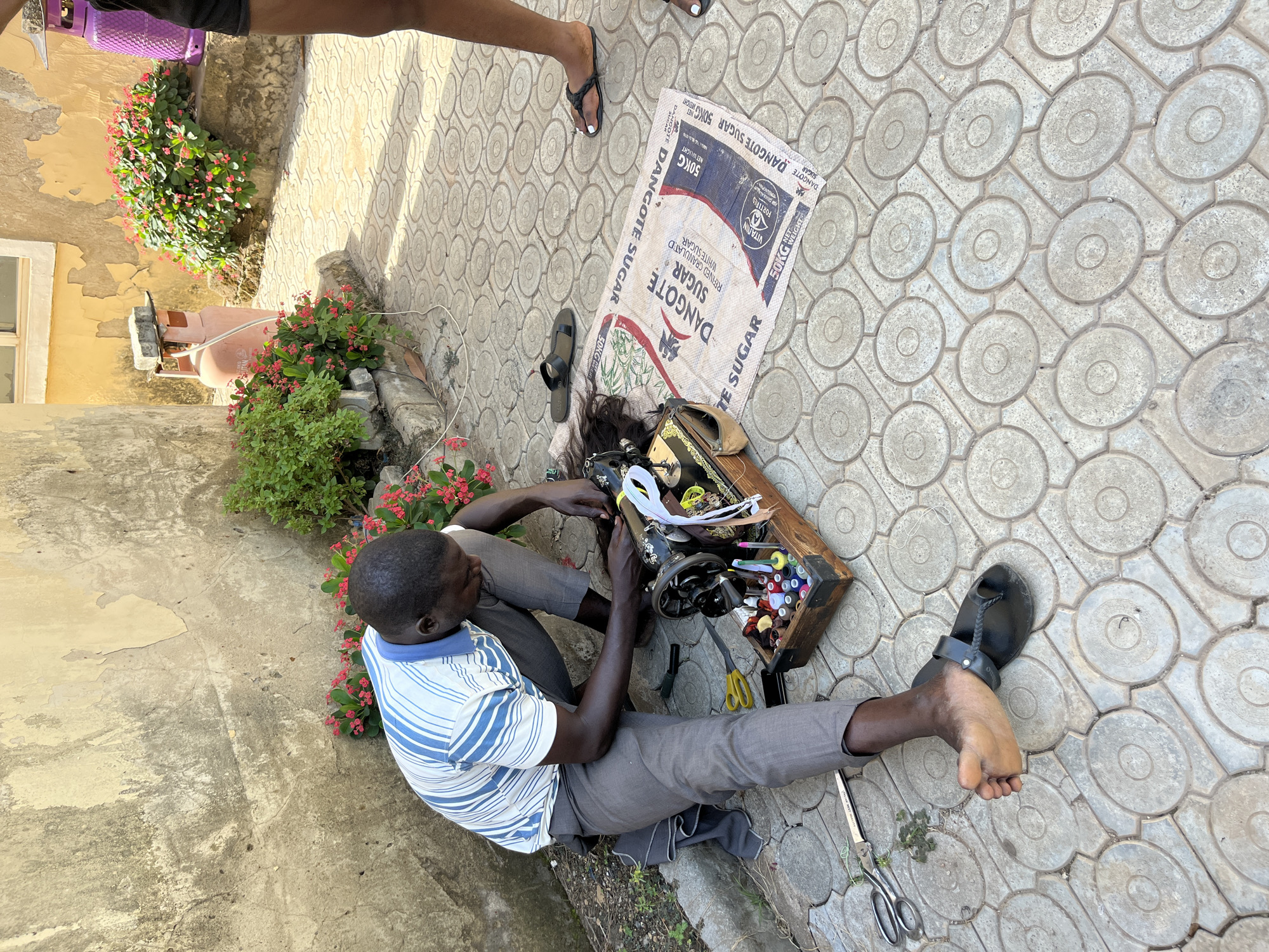 A man sits on a patterned pavement in an outdoor setting. He is using a manual sewing machine placed on a wooden box containing spools of thread and other sewing tools. Beside him is a discarded Dangote Sugar sack. The location is in Abuja, Nigeria. A building with a yellow wall and a patch of flowers are next to him. Two people in sandals are standing behind the sewer.