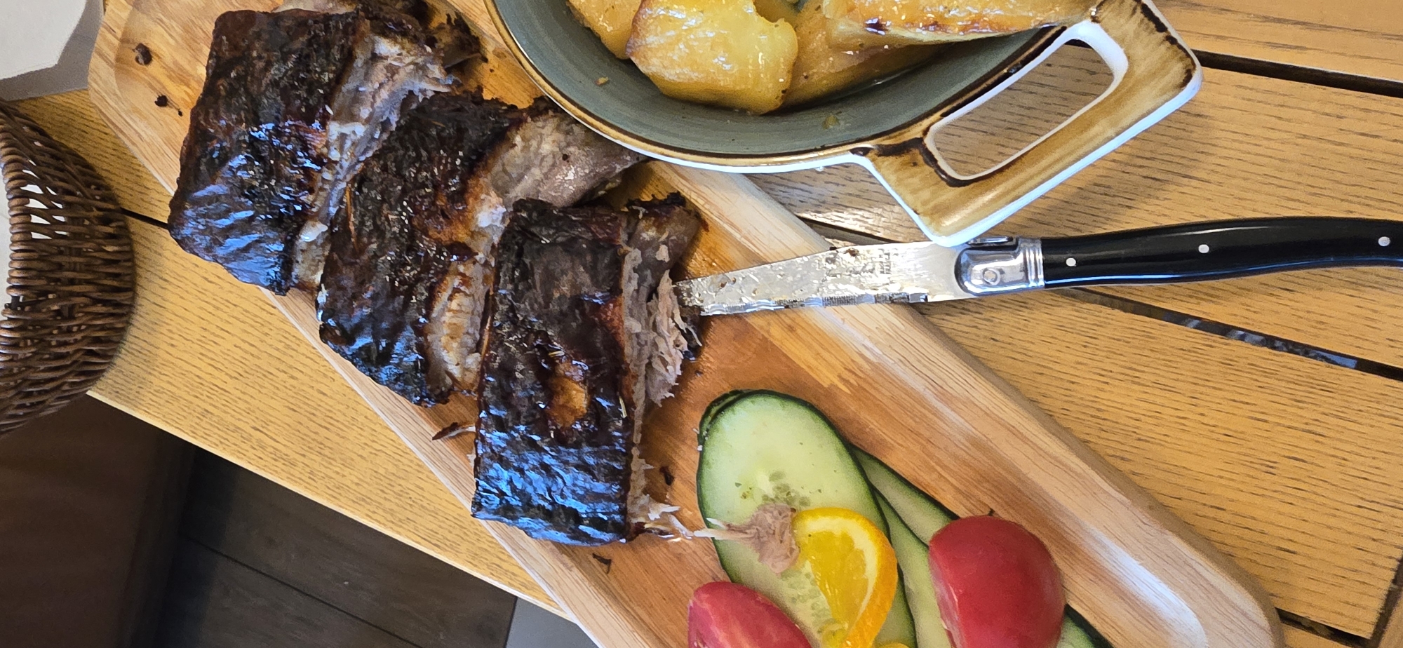 A high-angle shot shows a wooden tray with food on a wooden table. The tray holds ribs covered in a dark glaze, a dish of roasted potatoes in an abstractly shaped bowl, cucumber slices, tomato halves, and an orange slice. A black-handled Laguiole knife rests on the tray, partially cutting into the ribs. A woven basket sits on the table to the left. The location is Bigor, Montenegro. No people are visible.