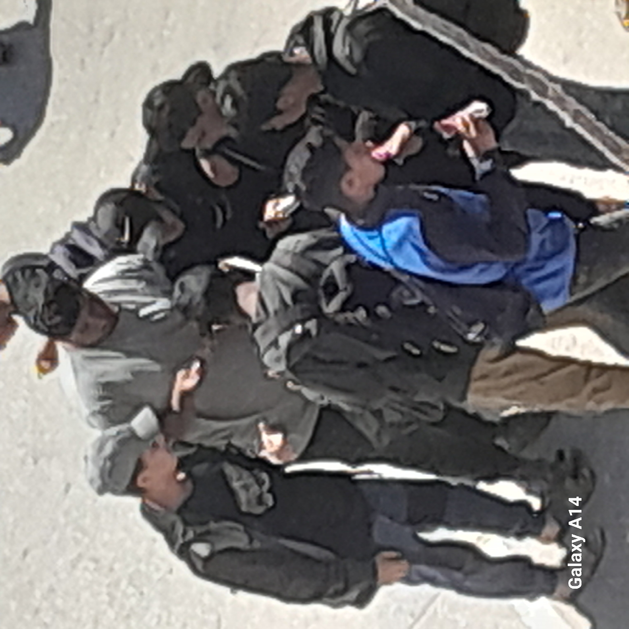The image displays a group of approximately seven to eight individuals, primarily adult males, gathered closely outdoors. They are standing on a light-colored ground, possibly pavement or concrete. Most of the subjects are dressed in dark jackets, hoodies, and casual clothing, with one person distinctively wearing a bright blue top. Several individuals appear to have backpacks or bags.
The group seems to be engaged in an activity or observing something together, as many are looking towards a common point, with some gazing upwards. One person, wearing the blue top, is holding a small, possibly reflective object to their mouth, suggesting they might be drinking or eating. Another individual, visible in the foreground with a grey cap, is also looking upwards.
The scene is brightly lit, indicative of daytime, with strong shadows cast on the ground, suggesting clear and sunny weather. There are no distinct architectural features or landmarks to identify the specific location, but it appears to be an urban or public outdoor setting. The visible text "Galaxy A14" is present as a watermark in the bottom right corner of the image. The emotions of the individuals are not clearly discernible due to the image quality and distance, but they appear focused and engaged.