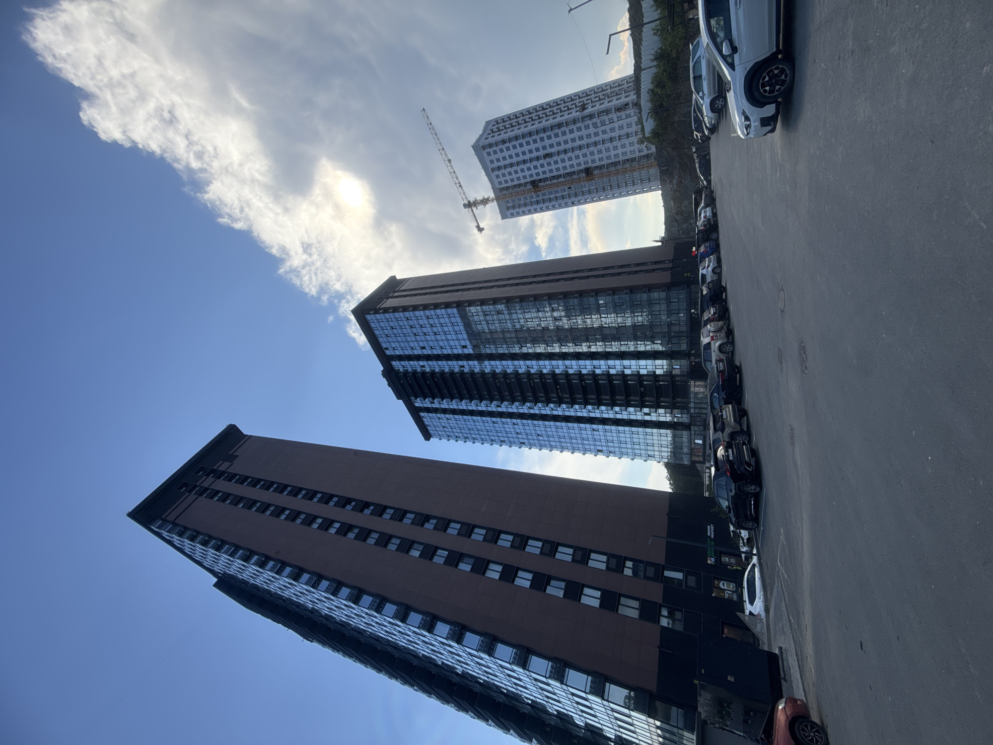 This is a low-angle, rotated shot of a modern cityscape with skyscrapers and a construction site. The foreground features a parking lot with several cars, including a white SUV, a dark sedan, and a red hatchback. The background is dominated by two large, contemporary buildings. The building on the left is taller and features a dark, reflective glass facade. To its right, a slightly shorter building has a reddish-brown facade with rows of windows. Further in the distance, a very tall, white building is under construction, with a prominent yellow crane extending from its side. The sky is a bright blue with scattered white clouds, and the sun is visible, suggesting it's daytime, possibly late afternoon given the warm light. The scene conveys a sense of urban development and modernity. The image appears to be taken in Vladivostok, Russia, as indicated by the location context. No people are visible in the frame. The text "ROBIN HOOD MARKET" is partially visible on a building facade in the lower-left portion of the image.