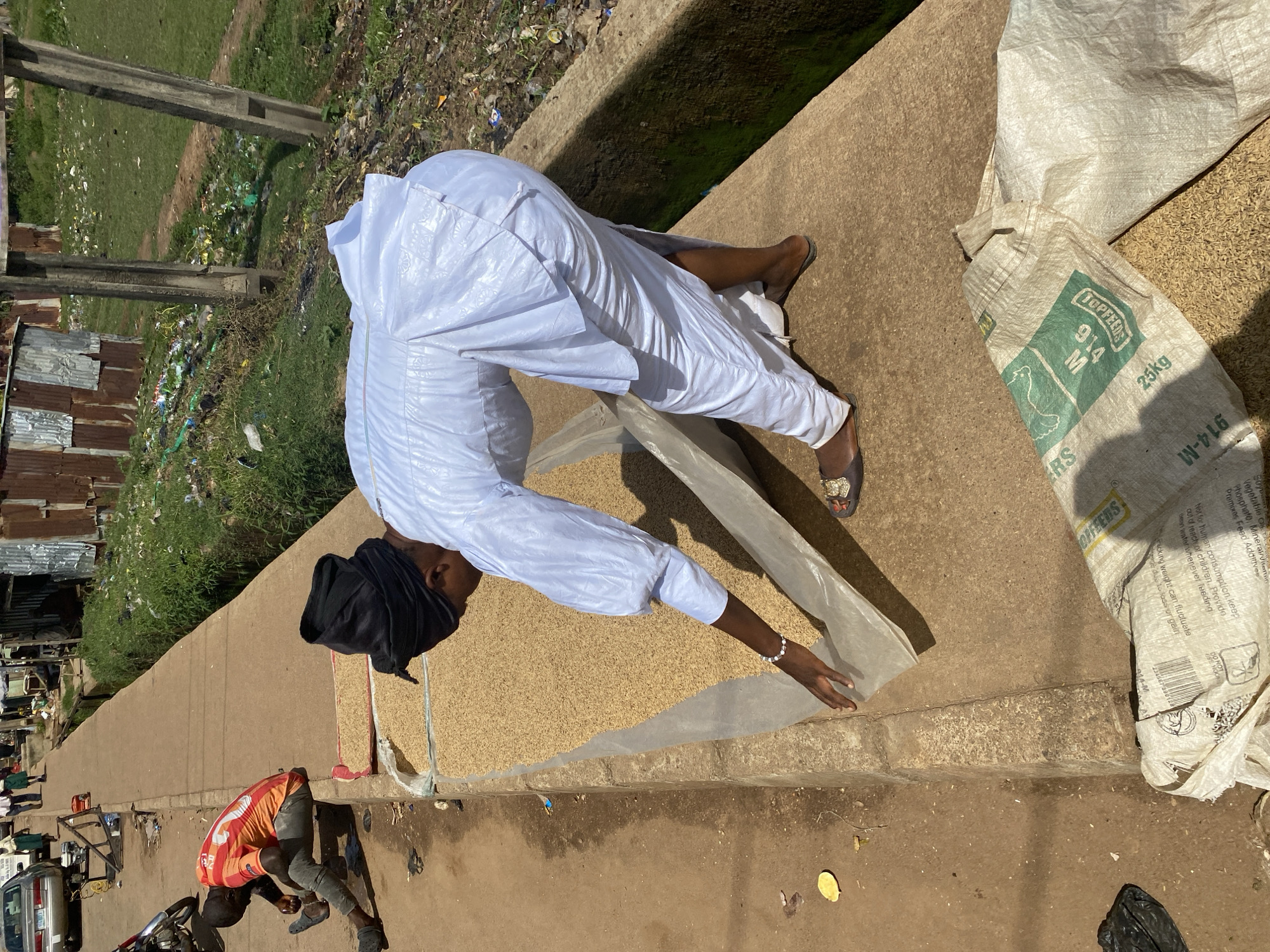 This media file captures a scene in Bukuru, Nigeria, depicting two individuals and bags of grain. The main subject is a person dressed in a white traditional outfit, kneeling and reaching towards a large spread of grain laid out on a plastic sheet. Their head is covered with a black cloth. To the right of the grain, there are two large white sacks. One sack, partially visible, is plain white, while the other, closer to the foreground, is a patterned bag with green and white text. Visible text on this sack includes "TOPFEEDS", "914 M", "25kg", and "914-M", along with nutritional information and warnings such as "Not for human consumption, keep out of reach of children."
In the background, another person is visible, wearing an orange and red striped shirt and grey pants, also bending down near a motorcycle. The setting appears to be an outdoor marketplace or roadside area. Evidence of urban life is present, with discarded items, corrugated metal structures, and a car visible in the distance. The bright sunlight suggests it is daytime. The overall scene suggests a commercial activity involving the handling of grains, likely for sale.