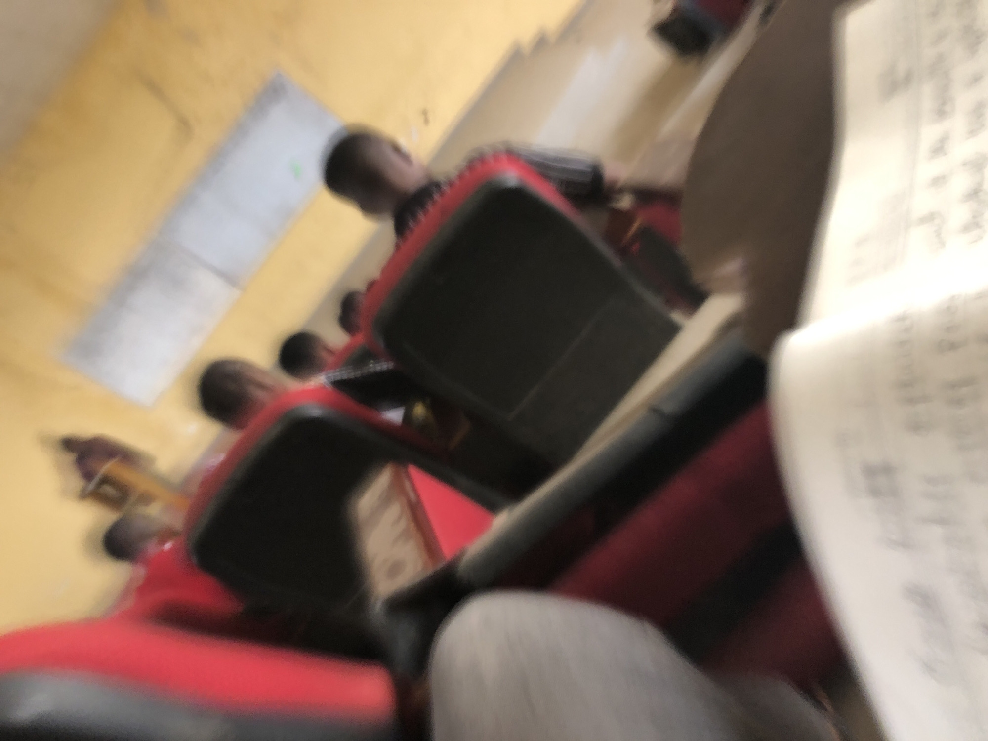 The image, likely taken in Pindiga, Nigeria, depicts a classroom setting. Several students are seated at desks with red chairs. A document with printed text is prominent in the foreground. The room has yellow walls and what appears to be a window. The scene appears blurred and taken from a low angle, possibly from the perspective of someone seated.