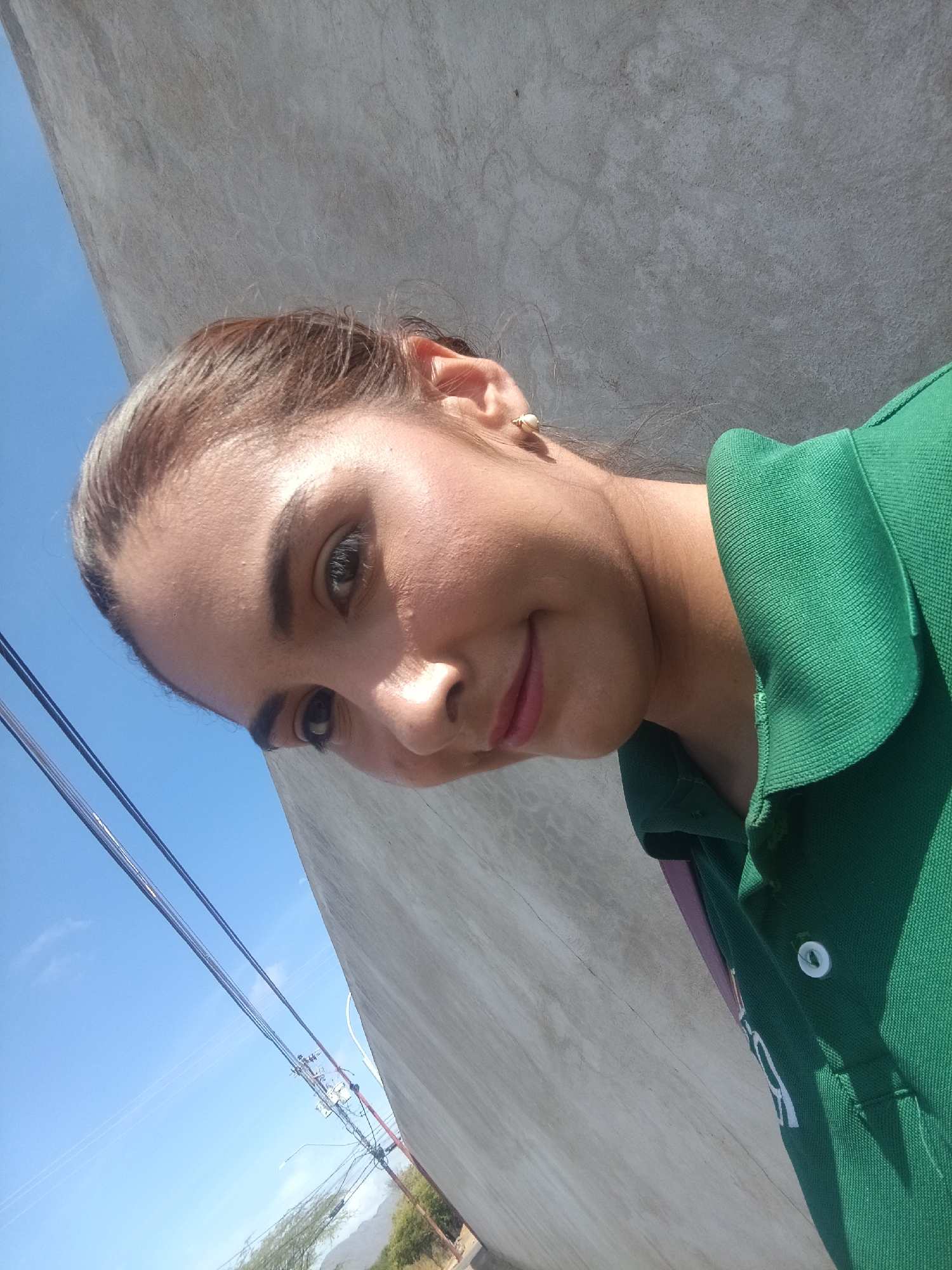A woman is outdoors in Juan Griego, Venezuela, standing near a concrete structure. The weather appears sunny, with a blue sky visible behind overhead power lines. She is wearing a green collared shirt with a white button. She wears a small, pearl-like earring. The background shows hints of vegetation and a distant hill or mountain. The wall is concrete, with visible cracks and variations in texture.