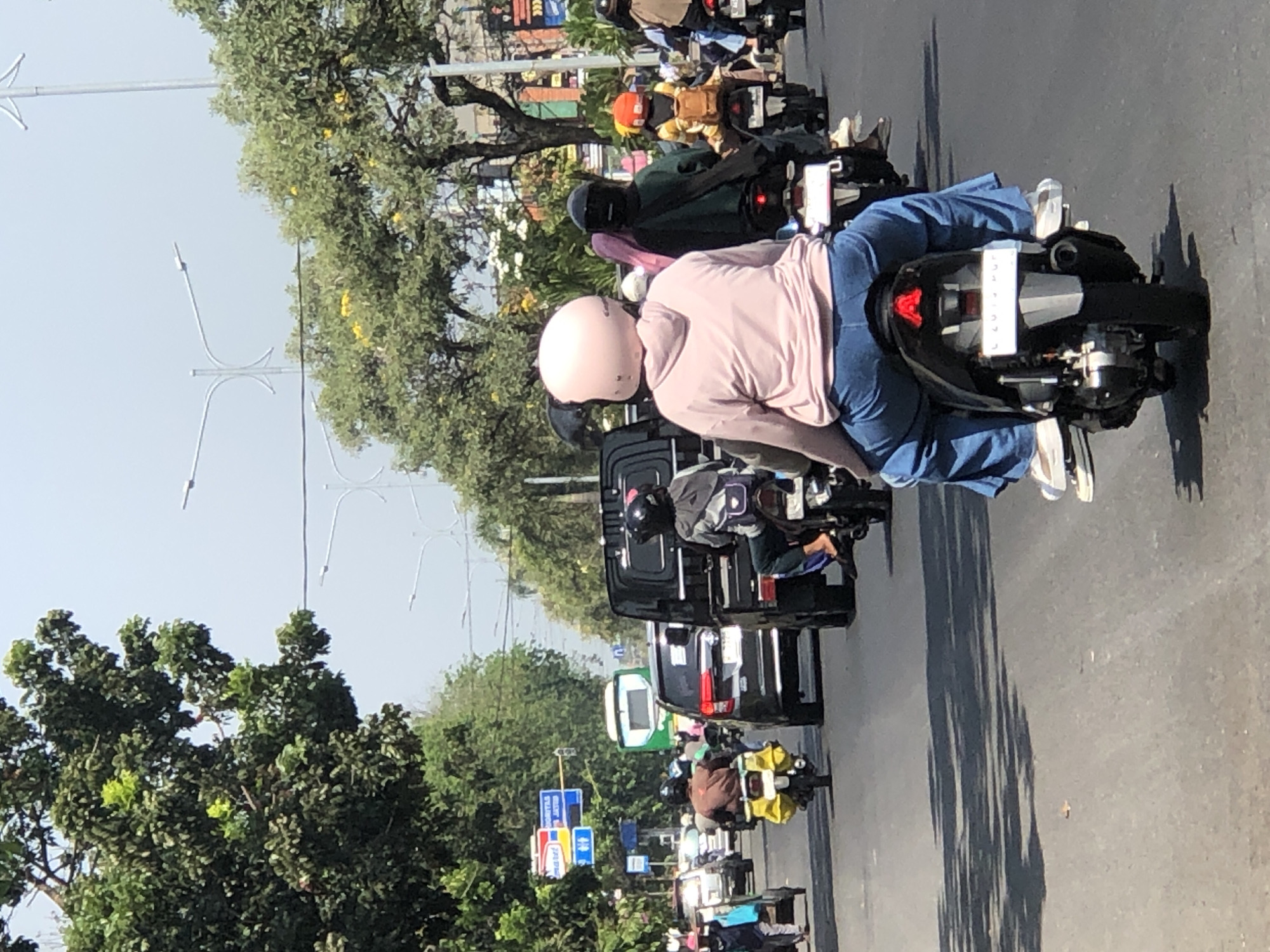 In Larangan, Indonesia, a busy street scene unfolds under a hazy sky. A procession of motorcycles navigates the asphalt, their riders and passengers clad in helmets and casual attire. The lead motorcyclist, facing away from the camera, wears a pink helmet and a light pink hooded sweatshirt, complemented by loose-fitting blue jeans. Behind them, other riders are visible, some with helmets of various colors, including black and orange, and others with headscarves. A green bus and a dark-colored SUV are also present in the traffic, along with a black pickup truck. The roadside is lined with lush green trees, some bearing yellow flowers, and streetlights stand tall against the pale sky. Signs, including one with blue and red markings and symbols for male and female restrooms, are visible on the left side of the road, indicating a public area. The overall impression is one of a typical day in a bustling Indonesian city.