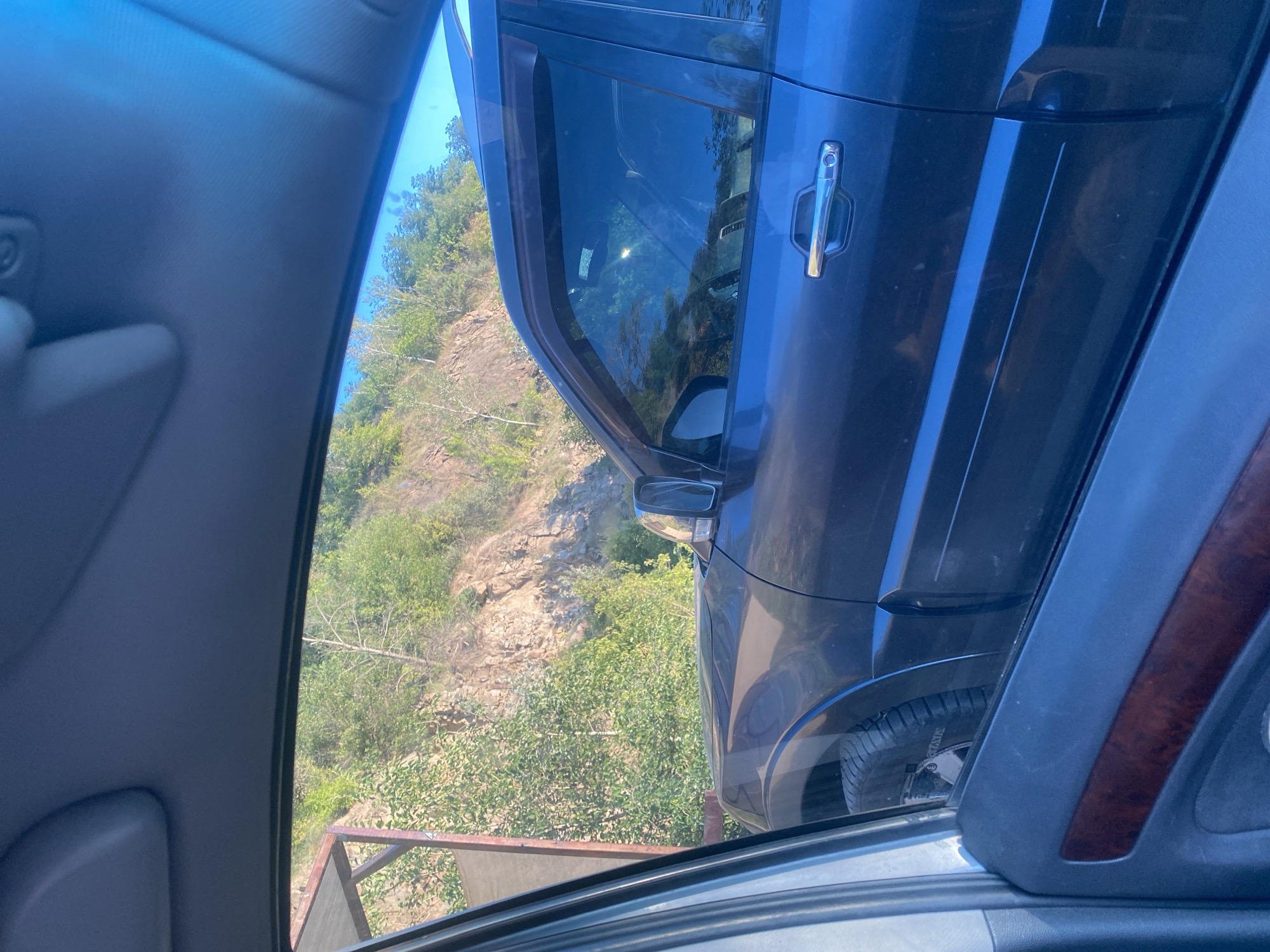 This image depicts the side of a dark blue SUV viewed from the interior of another vehicle, likely through the passenger window and side door. The SUV is parked on a road with a steep, rocky, and vegetated hillside to its right. The scene appears to be in an outdoor, possibly rural or semi-rural, environment. There are no people visible. The sun is shining, casting reflections on the SUV's windows. The interior of the viewing vehicle shows a portion of its door panel with wood trim and the roof liner. The image does not contain specific landmarks or visual cues that would allow for a precise identification of its location within the city of Dnipro, Ukraine.