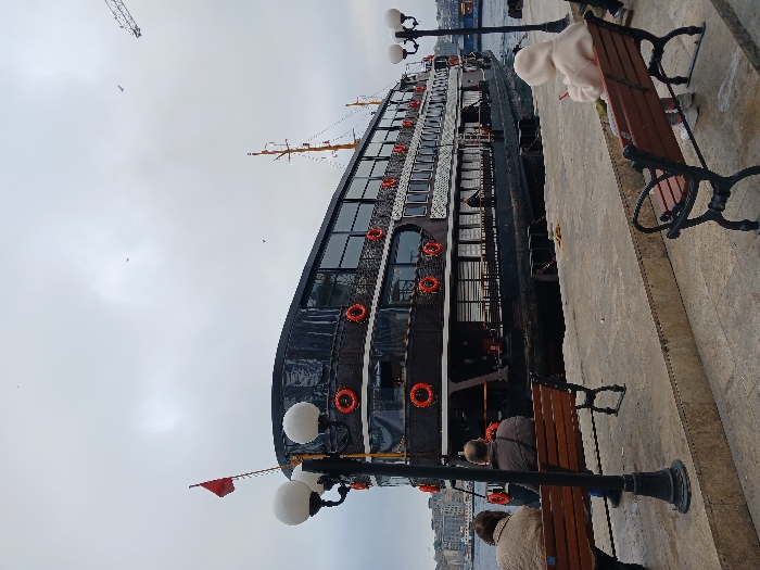 The scene takes place on a pier in İstanbul, Türkiye, near the coast, at the coordinates 41.022° N, 28.976° E. A black ferry boat is docked at the pier. There are three people in the scene: two sitting on a bench by a lamppost in the foreground and one seated on a bench in the lower right corner of the image. The two figures in the foreground are facing each other, and the third figure is facing the ferry boat. The scene is overcast and the sky is gray. The ferry boat is multi-level and has red life preservers around the railings. There is a small red flag flying from a pole at the top of the pier.