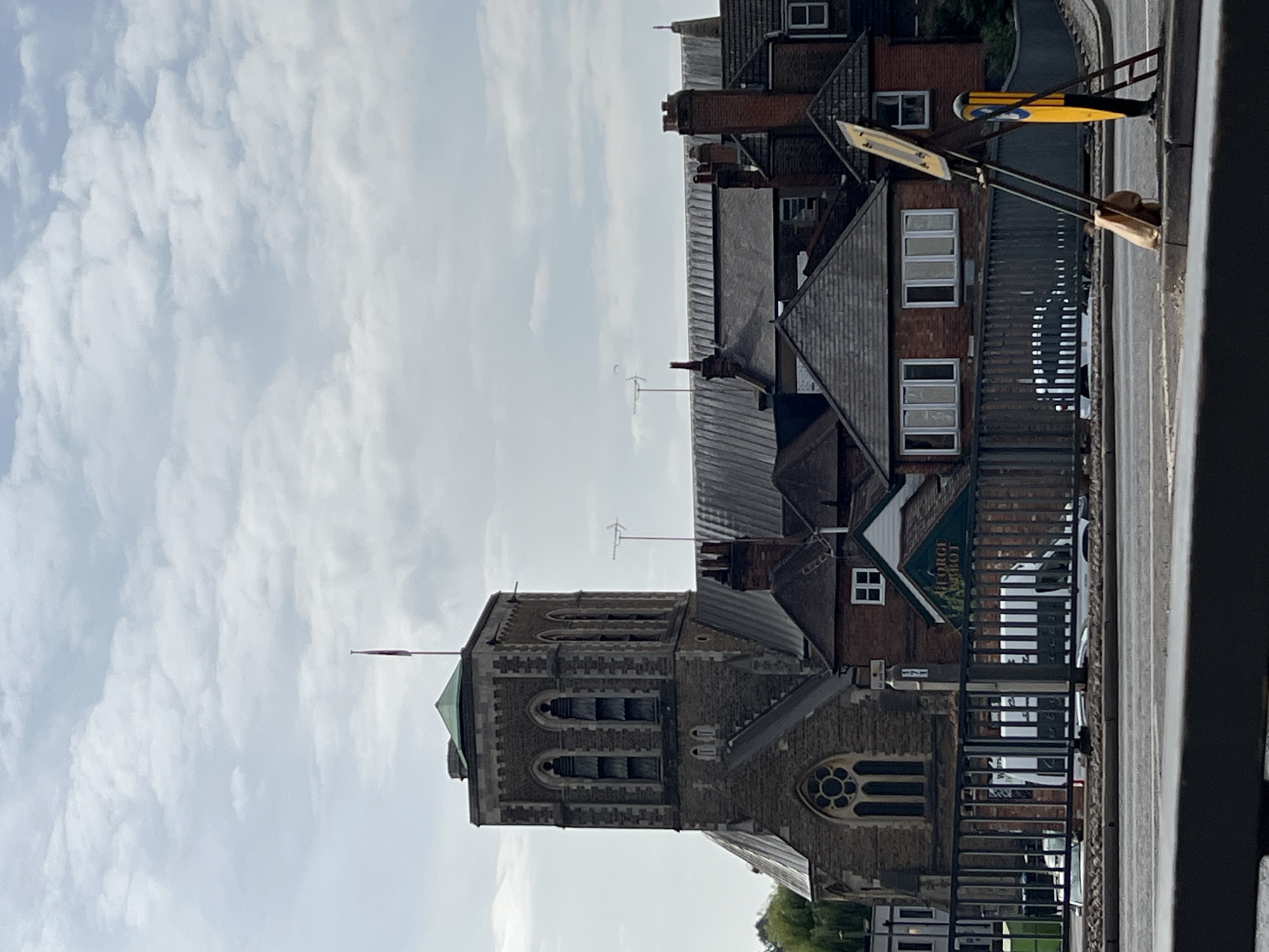 The image shows a street scene in Guildford, United Kingdom, featuring a large stone church with a prominent bell tower. The church has a green, conical roof at the top of the tower, and a flag is flying from a pole. The tower has arched windows with louvres. Adjacent to the church are several brick buildings with pitched roofs and chimneys. A metal fence runs across the foreground, behind which can be seen a parked white van and other vehicles. A yellow temporary road sign is propped up on the pavement. The sky is partly cloudy.