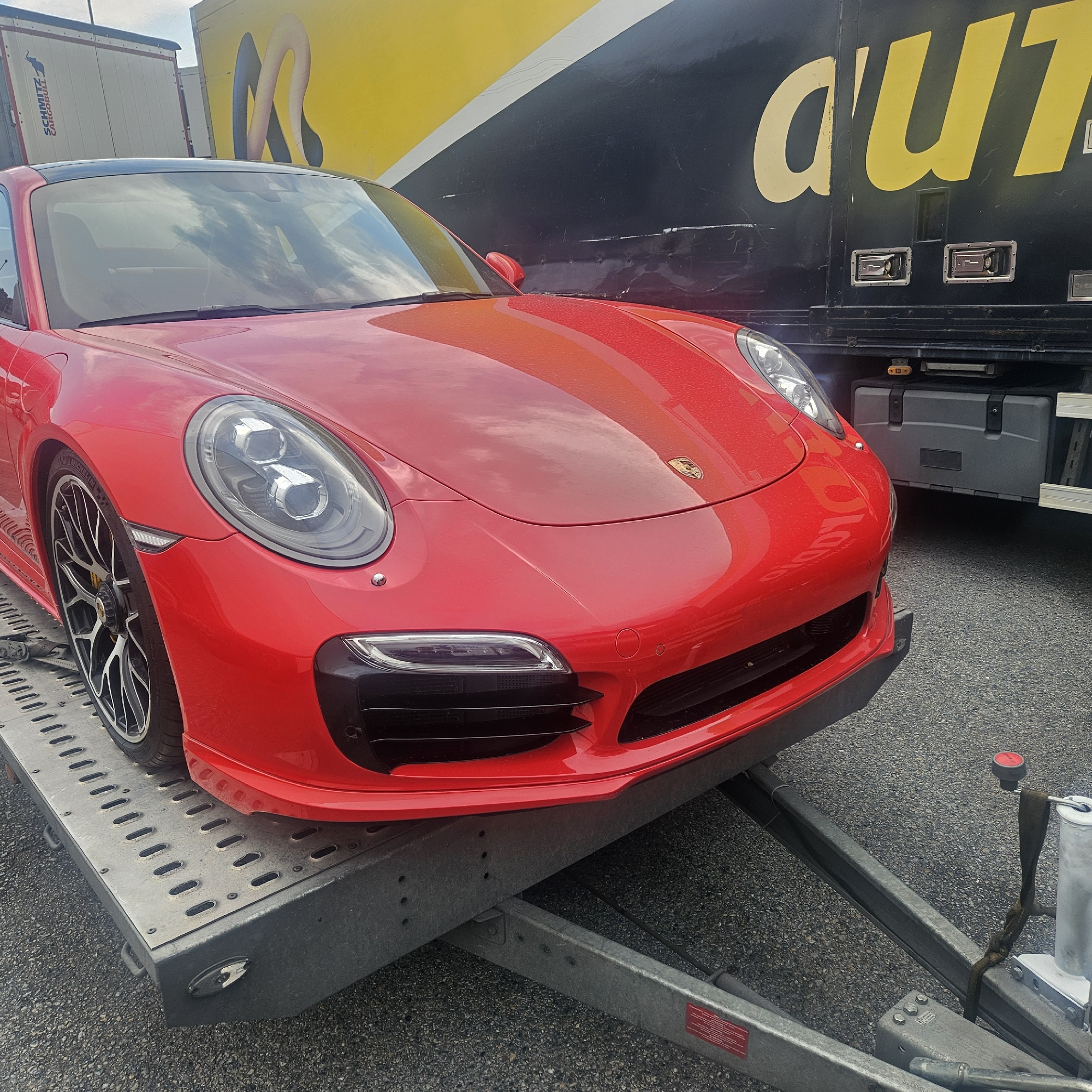 A bright red Porsche sports car is parked on a metal trailer, which is positioned on a grey asphalt surface. The car's front is angled towards the right side of the frame, revealing its sleek design, including its headlights, wheels, and a portion of its hood. The Porsche has a black roof. Behind the car, a large, dark-colored truck is visible, with a prominent yellow graphic on its side that reads "d'aut...". The truck appears to be parked in a lot, with a smaller white trailer with "SCHMITZ CARGOBULL" printed on it partially visible to the left. The scene is outdoors, with some reflections of clouds visible on the car's surface, suggesting an overcast or partly cloudy day. The overall impression is of a vehicle being transported or prepared for transport, likely in an industrial or commercial area.